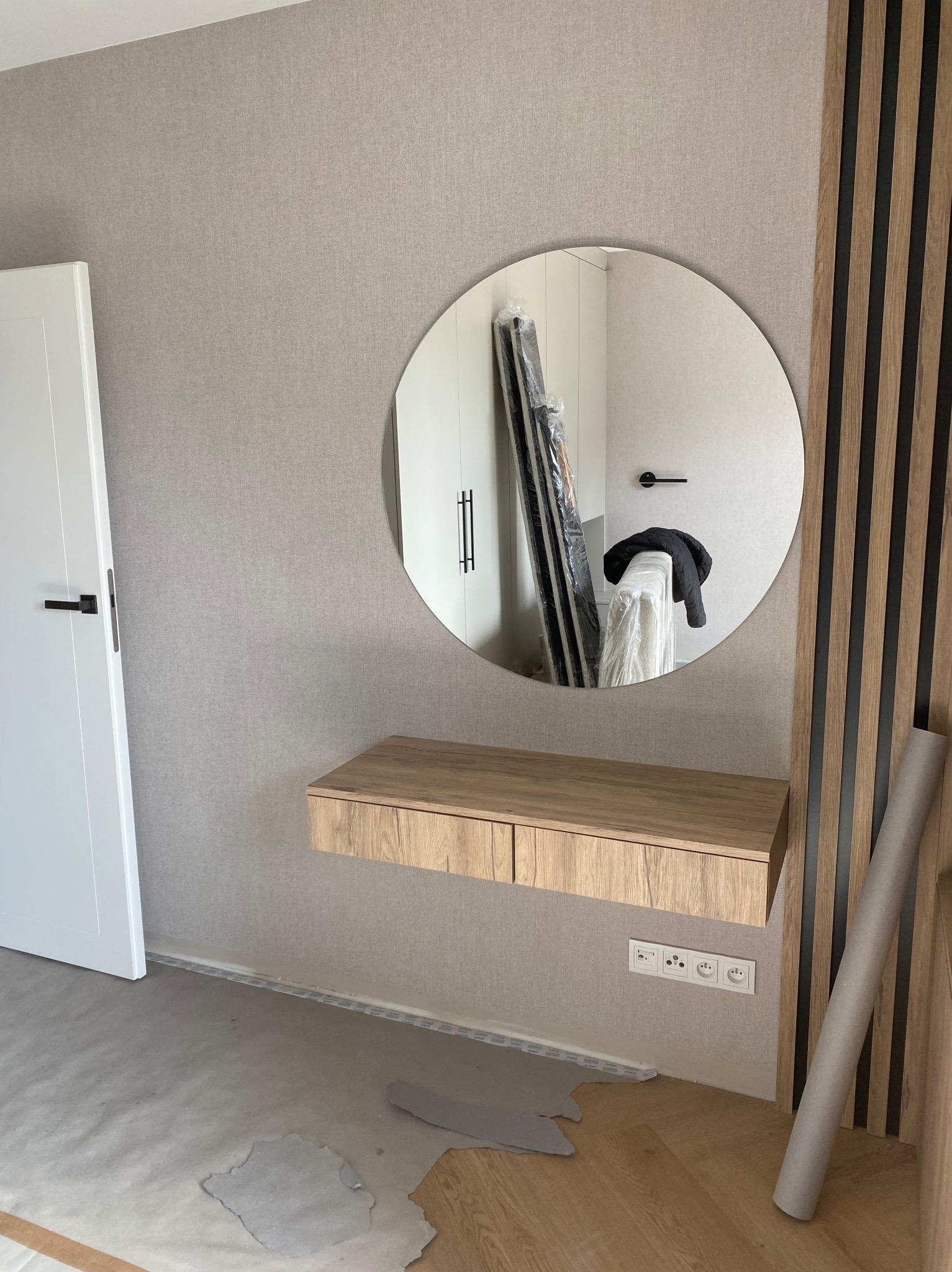 Okrągłe lustro nad drewnianą konsolą z szufladami, odbijające wnętrze z szafą. Dekoracyjne panele ścienne z drewna i czarnych listew po prawej stronie.