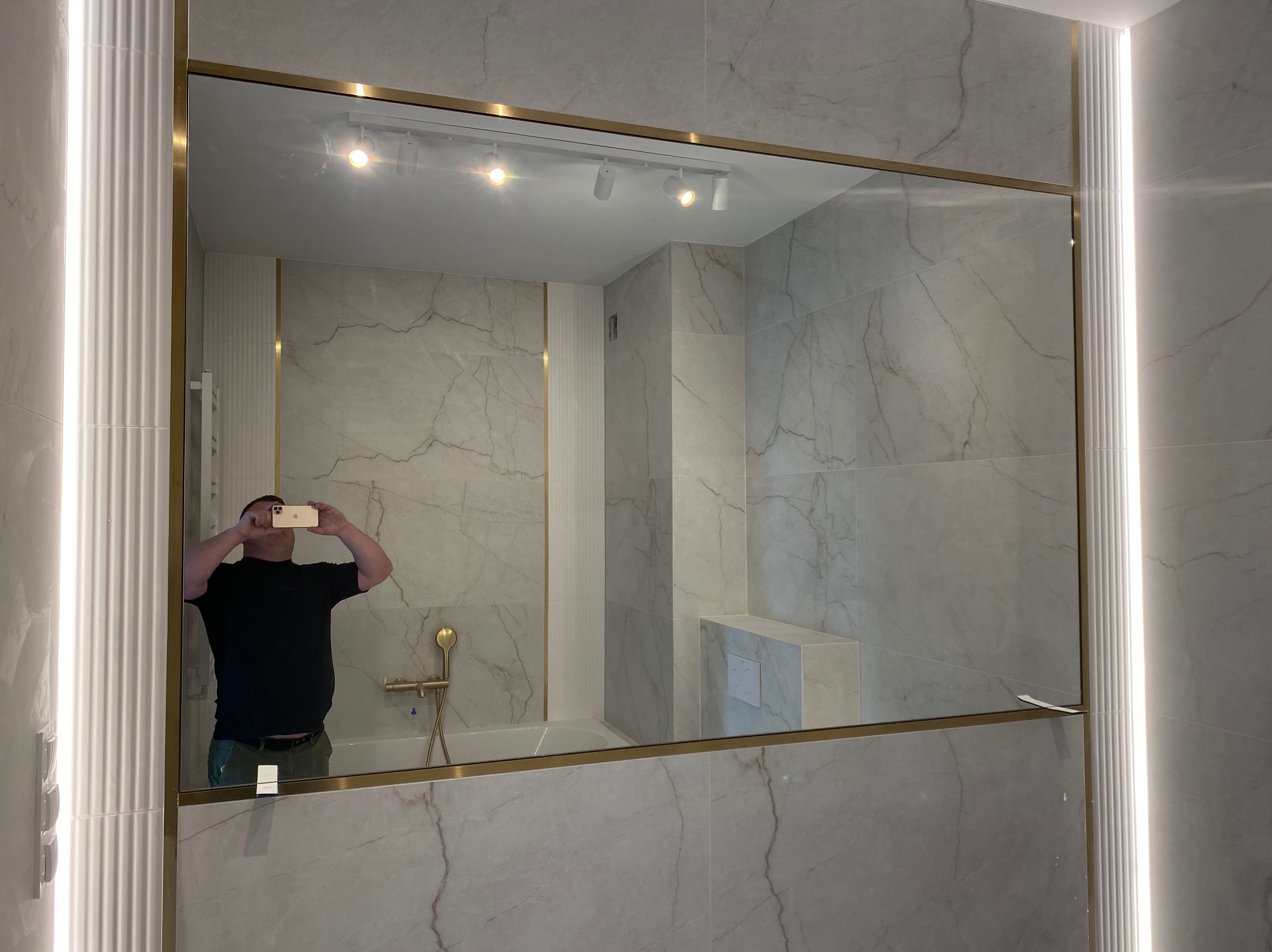 Lustro w złotej ramie w łazience z marmurowymi płytkami, odbijające wnętrze i osobę robiącą zdjęcie telefonem. Oświetlenie LED po bokach lustra.