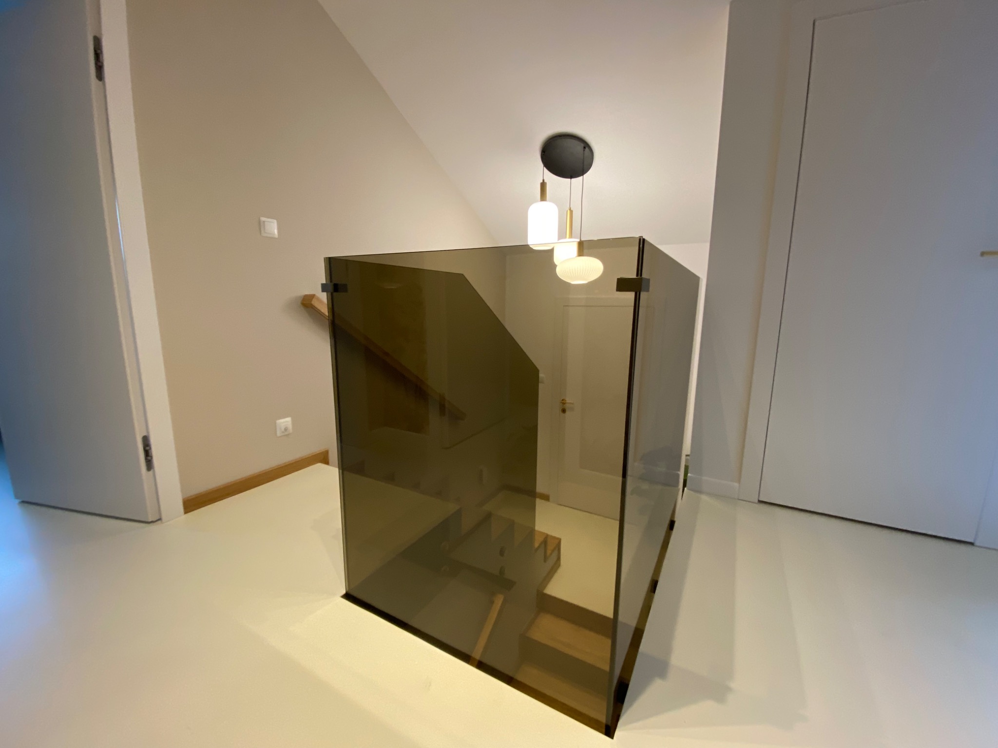 Szklana balustrada w nowoczesnym wnętrzu, widok na schody. Minimalistyczny design, neutralna kolorystyka ścian i podłogi, wisząca lampa nad balustradą.