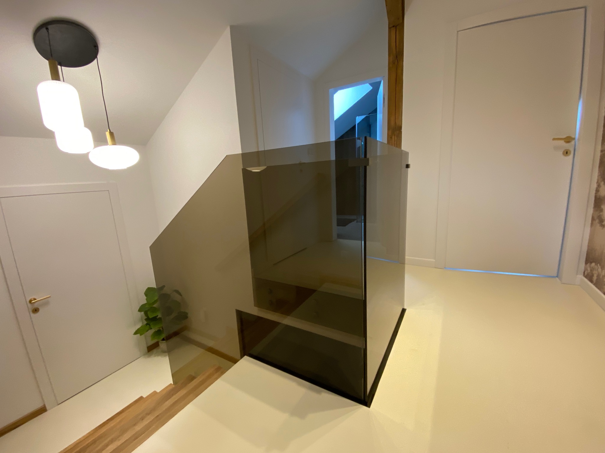 Szklana balustrada schodowa w nowoczesnym wnętrzu. Minimalistyczny design, neutralna kolorystyka, drewniane schody i białe drzwi. Widok z dołu, oświetlenie sufitowe.