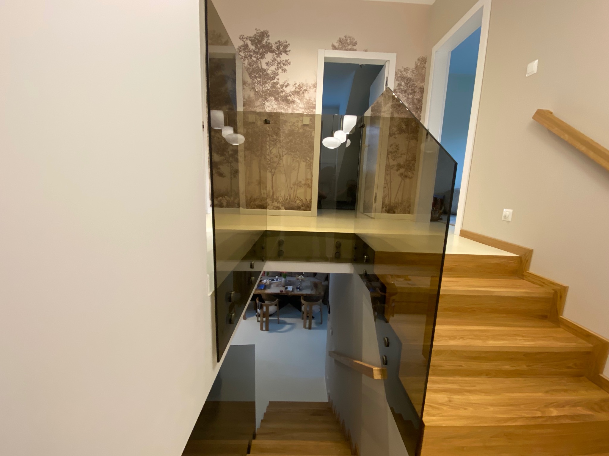 Nowoczesna, brązowa balustrada ze szkła na schodach z drewna, widok z góry. W tle tapeta z motywem drzew i widoczny fragment jadalni.