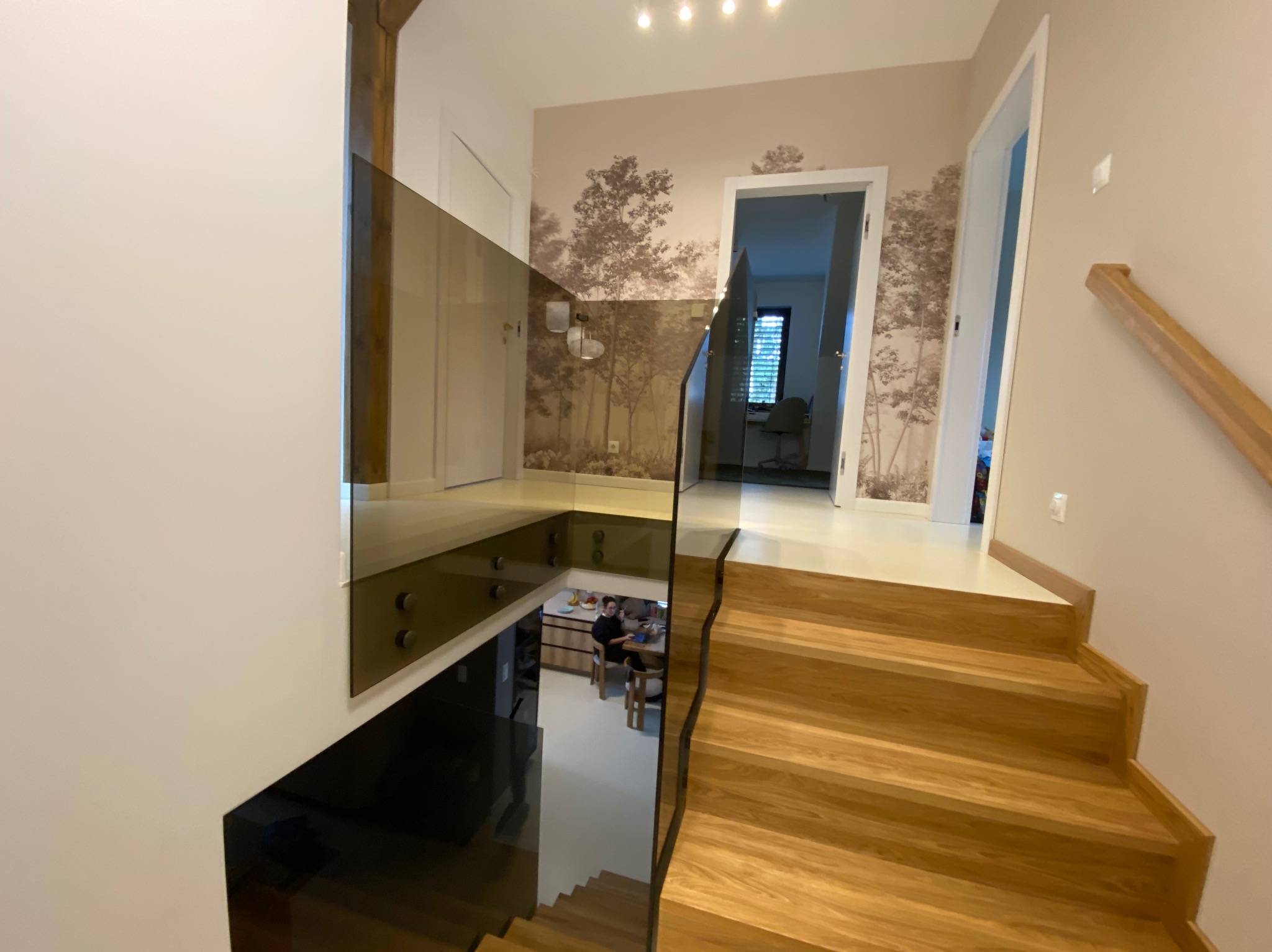 Nowoczesna, brązowa balustrada szklana na schodach z drewna, z widokiem na dół. W tle tapeta z motywem drzew i otwarte drzwi.