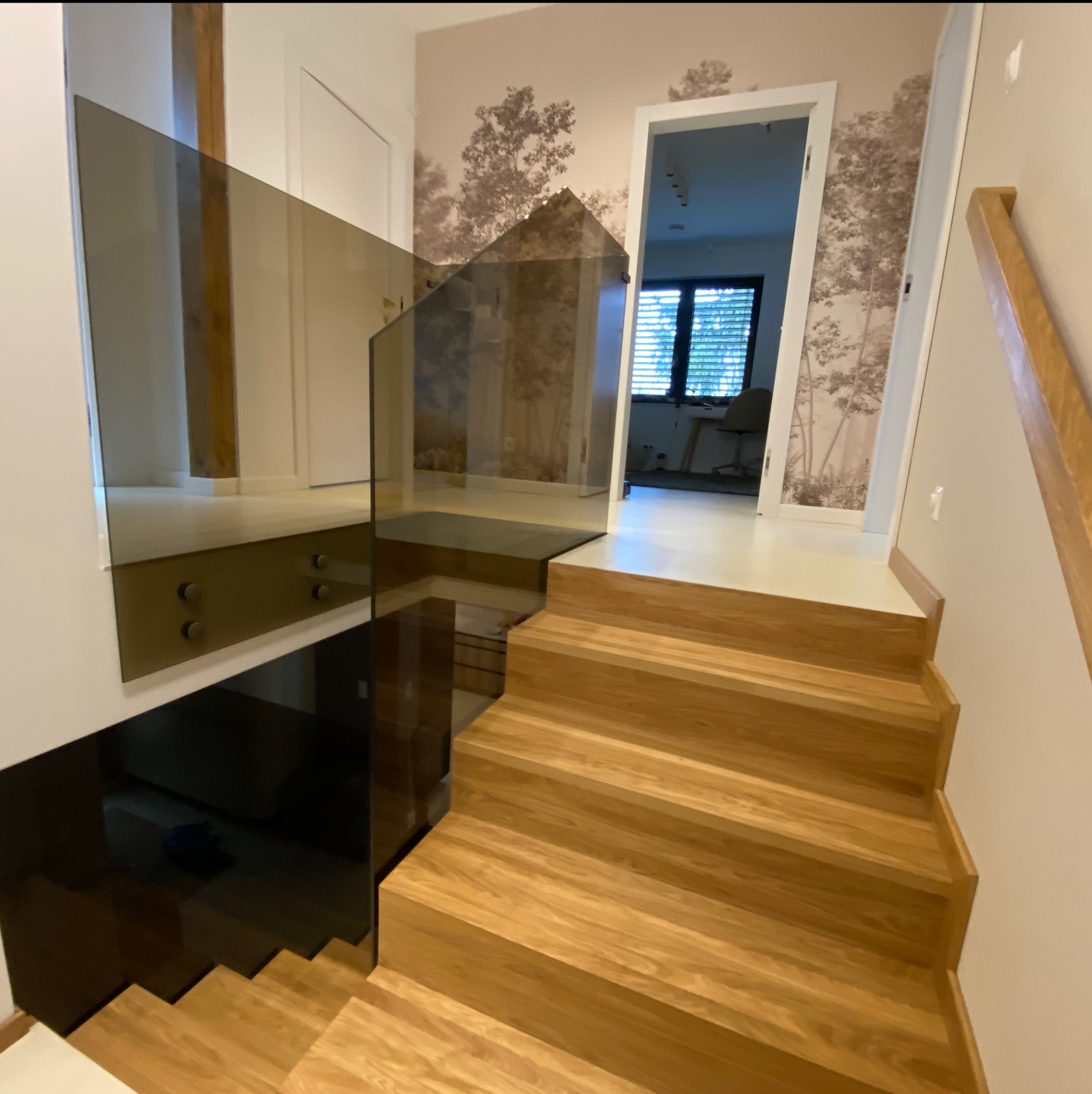 Nowoczesne schody z drewnianymi stopniami i szklaną balustradą, mocowaną na metalowych uchwytach. W tle tapeta z motywem drzew i otwarte drzwi do innego pomieszczenia.