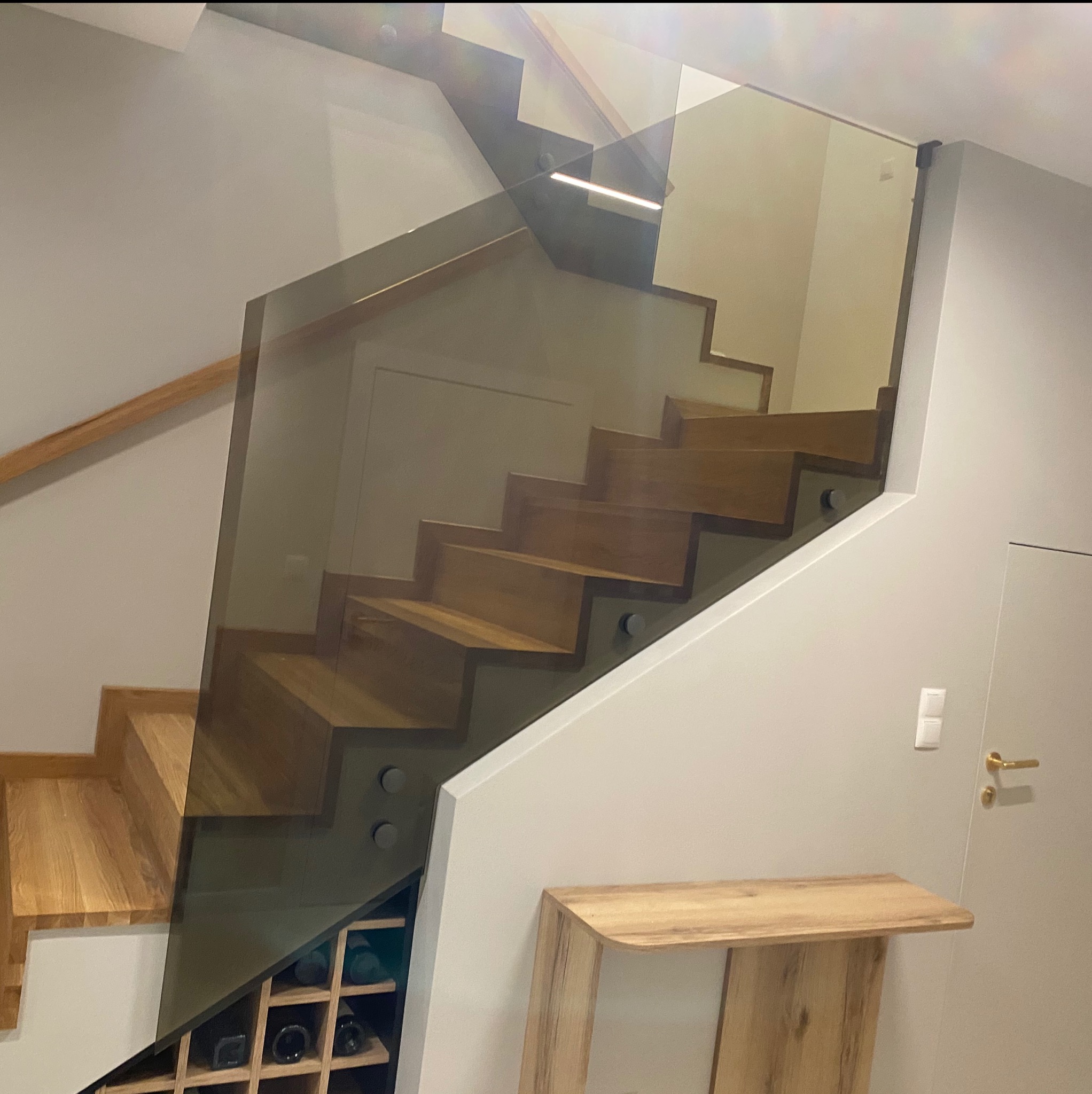 Drewniane schody z nowoczesną, szklaną balustradą mocowaną na metalowych elementach. W tle widoczna ściana i fragment półki.