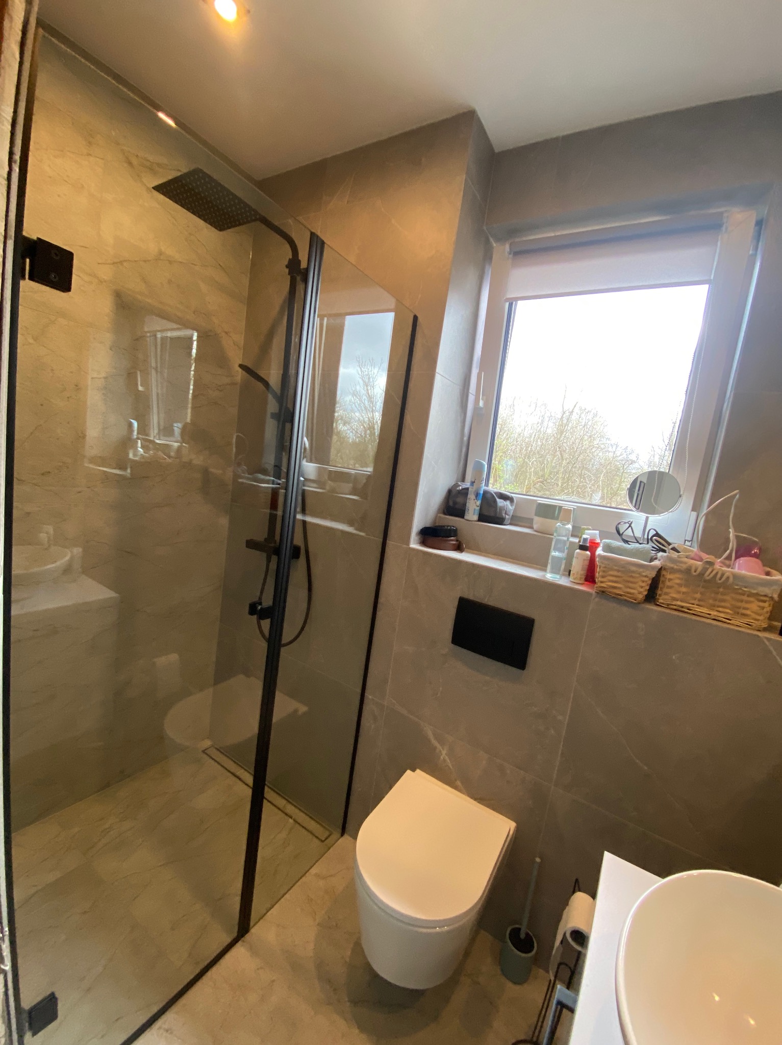 Nowoczesna kabina prysznicowa ze szklanymi drzwiami i czarnymi profilami w łazience wykończonej płytkami imitującymi marmur. Widok na okno z roślinnością.