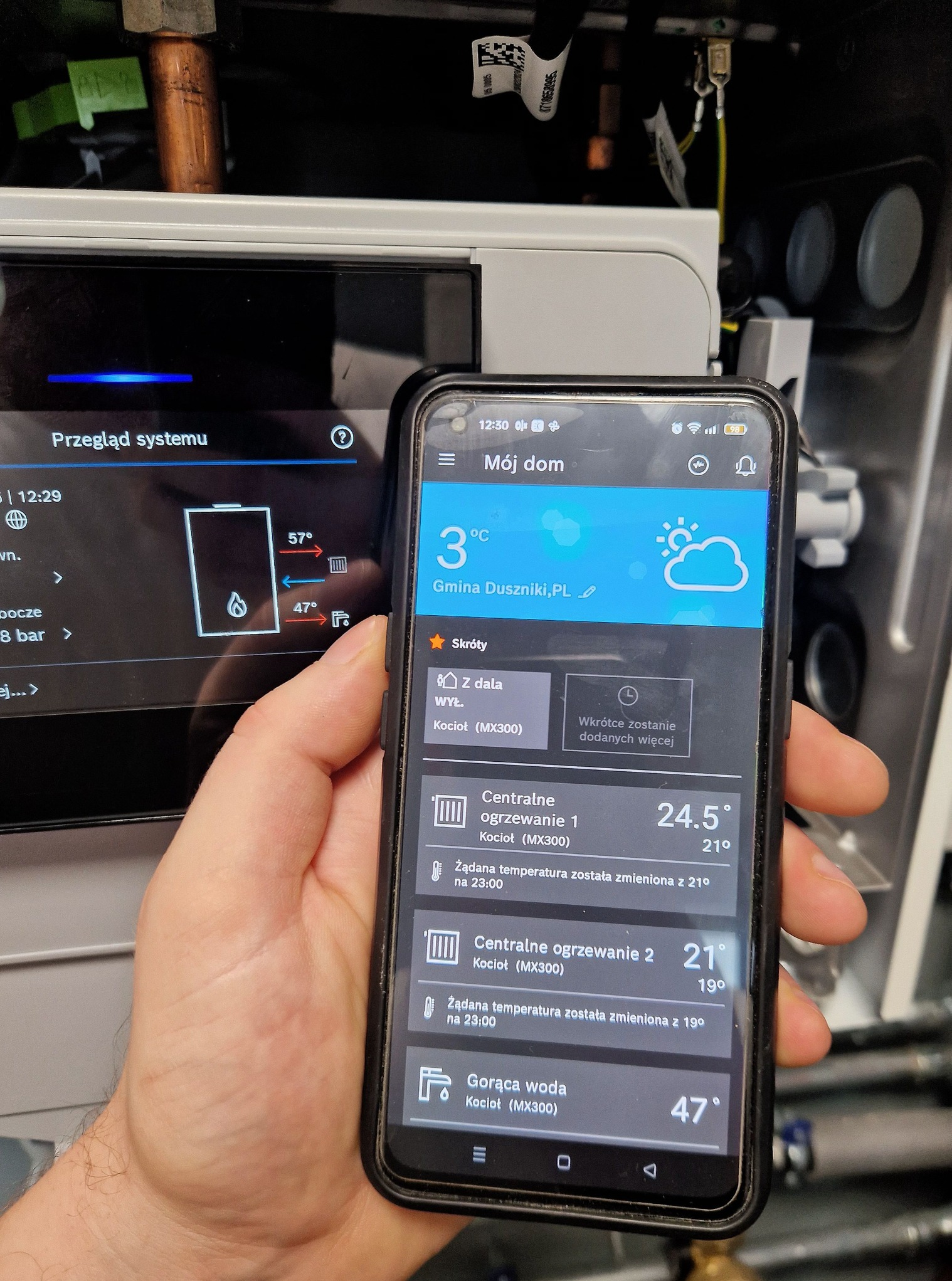 Smartfon z aplikacją do sterowania ogrzewaniem w dłoni, na tle panelu sterowania pieca. Widoczne ustawienia temperatury i status kotła w Gminie Duszniki.