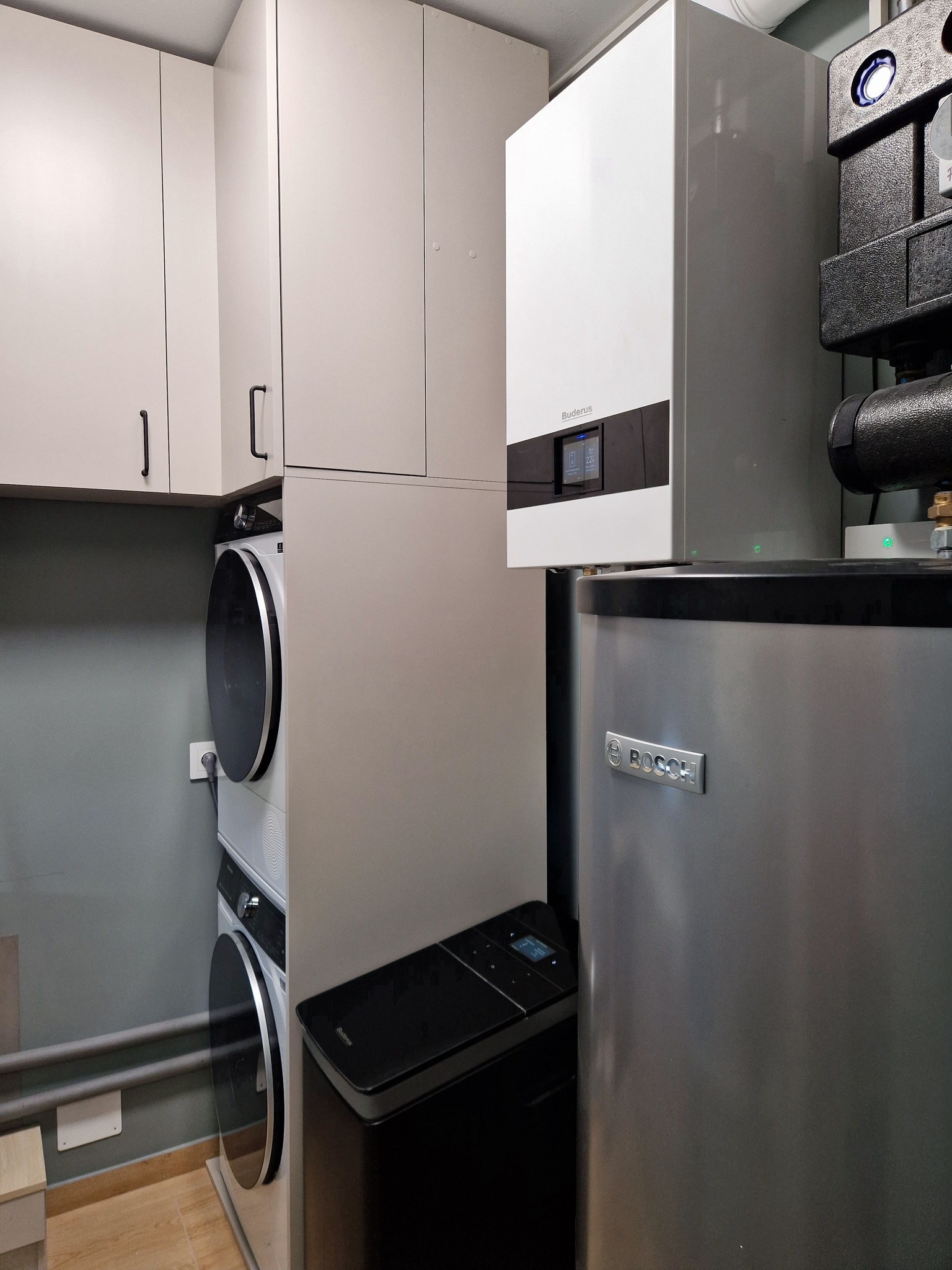 Kompaktowa kotłownia z piecem gazowym Buderus i zasobnikiem Bosch, obok pralka i suszarka w zabudowie. Szare ściany, beżowe szafki wiszące.