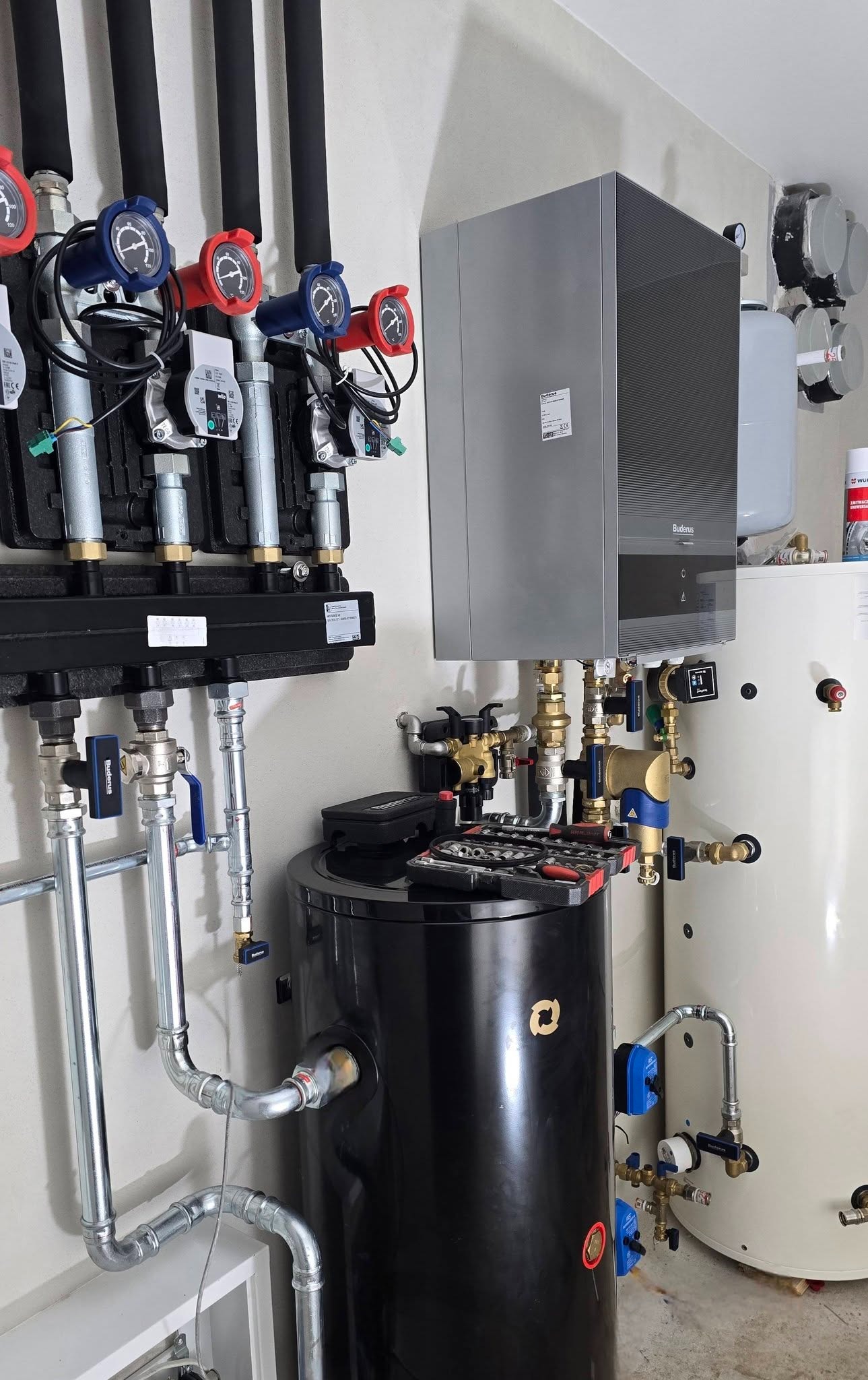 Nowoczesny piec gazowy Buderus z czarnym zasobnikiem i rozbudowaną instalacją hydrauliczną, widoczne manometry i zawory. Na zasobniku leżą narzędzia.