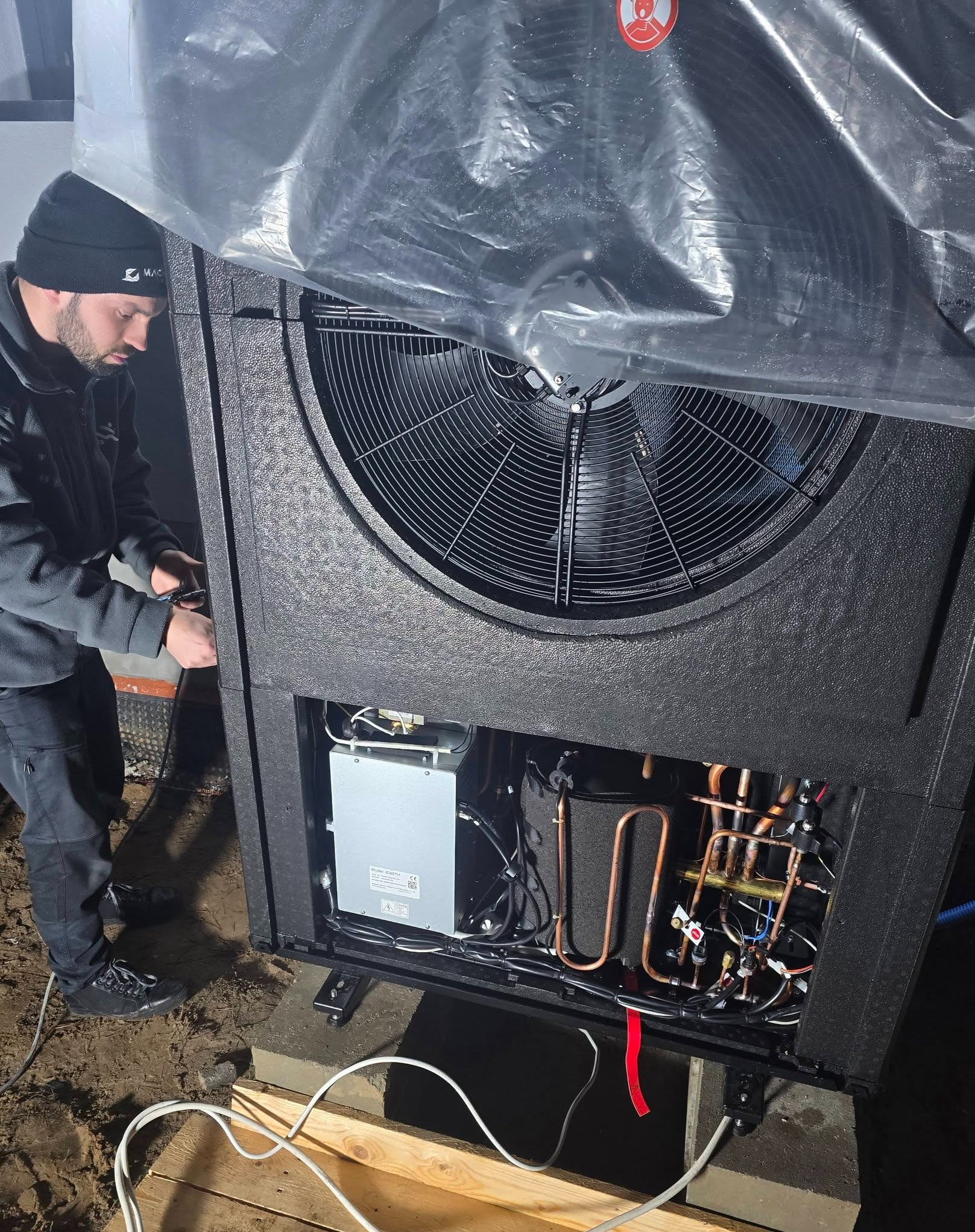 Instalator w czapce montuje pompę ciepła na zewnątrz budynku. Widoczne miedziane rury, wentylator i elementy elektryczne. Część urządzenia zakryta folią ochronną.