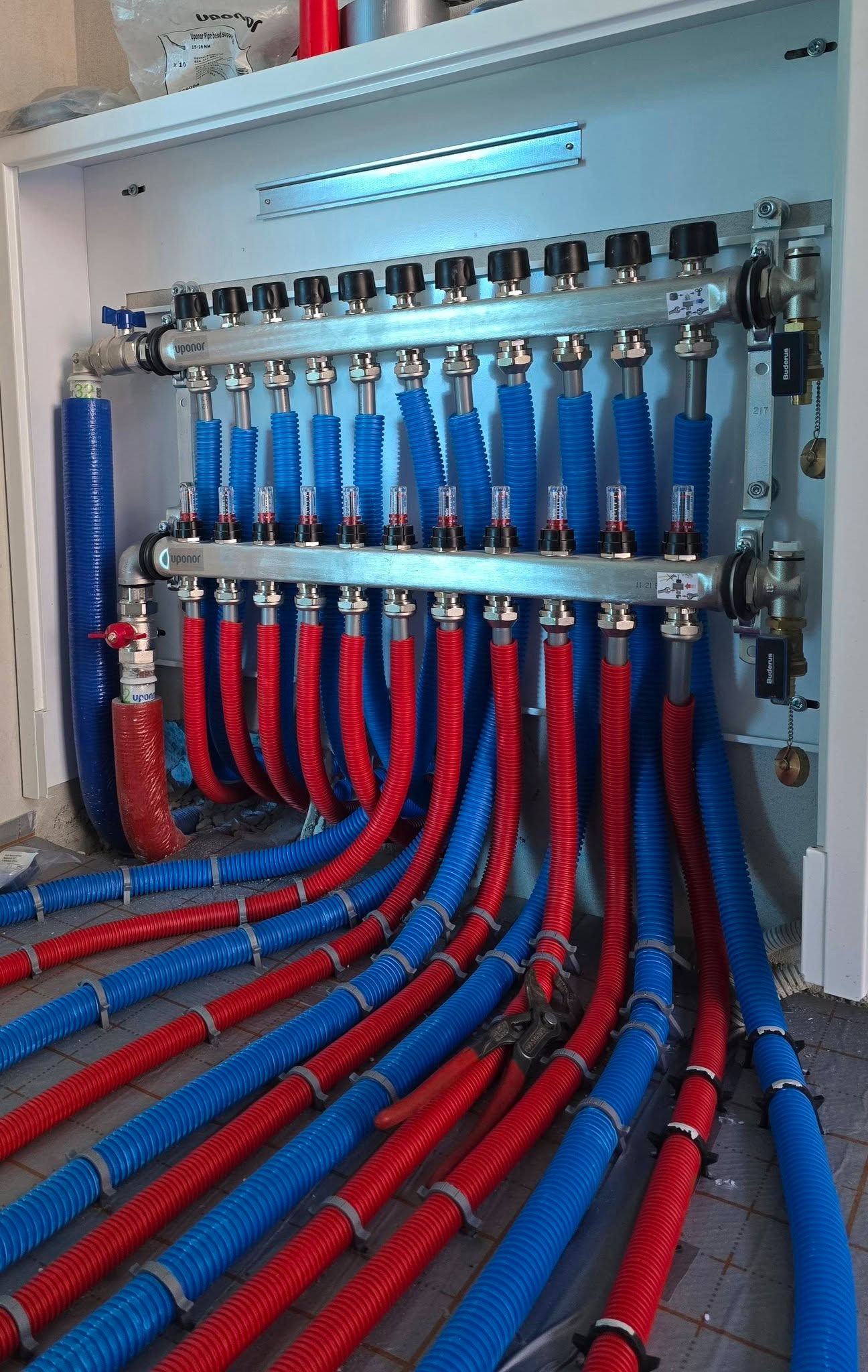 Rozdzielacz ogrzewania podłogowego Uponor z niebieskimi i czerwonymi rurami, zamontowany w szafce. Widoczne narzędzia i elementy instalacji.
