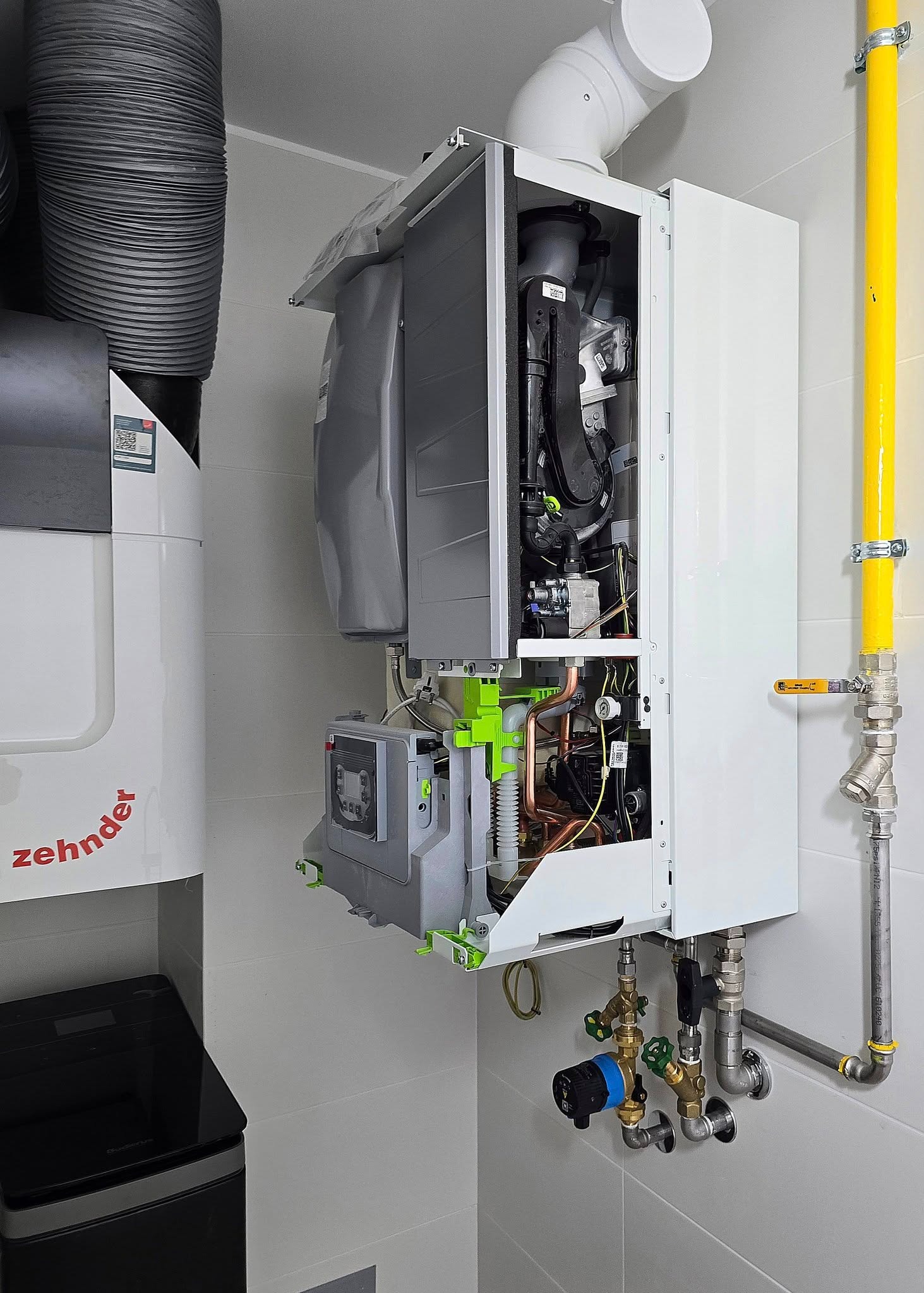 Otwarty, biały piec gazowy z widocznymi elementami wewnętrznymi i żółtą rurą gazową, zamontowany na jasnej ścianie z płytkami. Widoczny zbiornik Zehnder.