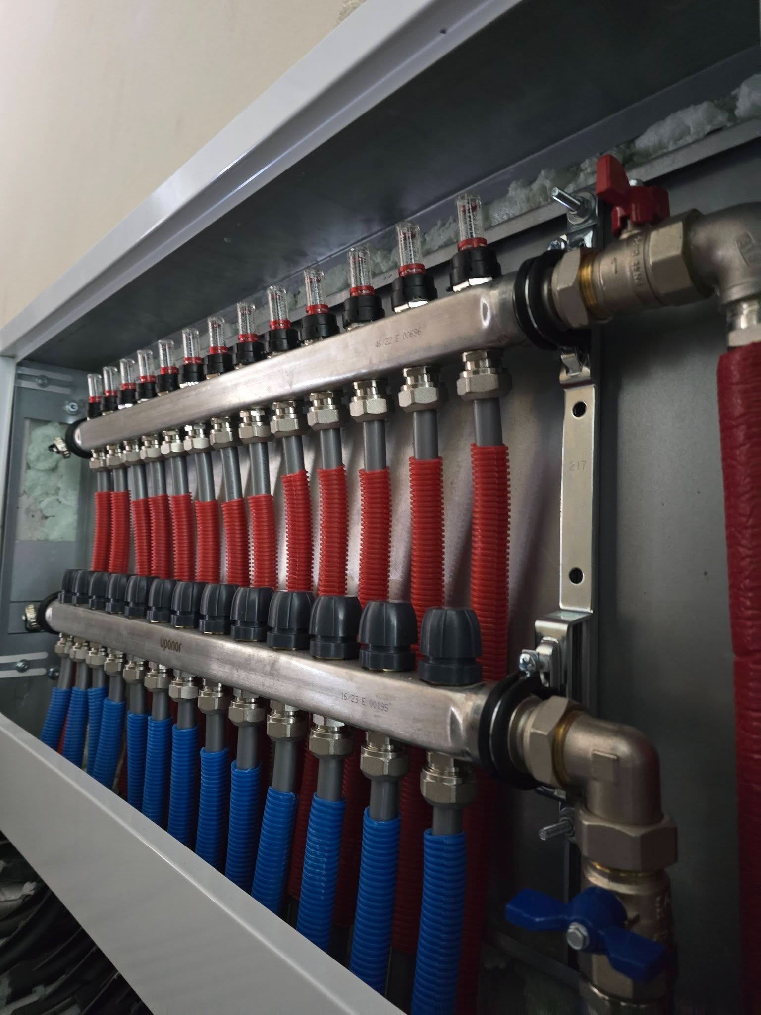 Rozdzielacz ogrzewania podłogowego Uponor z rurami w kolorze niebieskim i czerwonym, zamontowany w szarej szafce. Widoczne zawory i elementy instalacji.