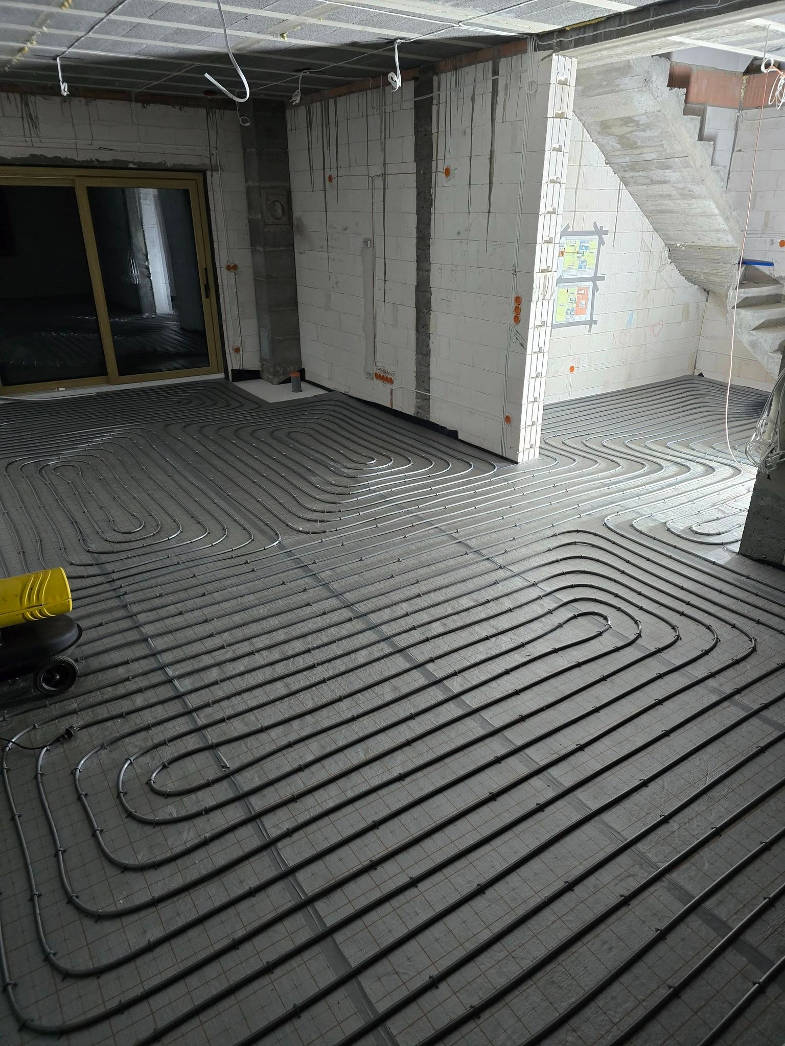 Szare rury ogrzewania podłogowego ułożone w pętle na podkładzie, widoczne ściany z betonu komórkowego i schody w surowym stanie. Żółty element maszyny w lewym dolnym rogu.