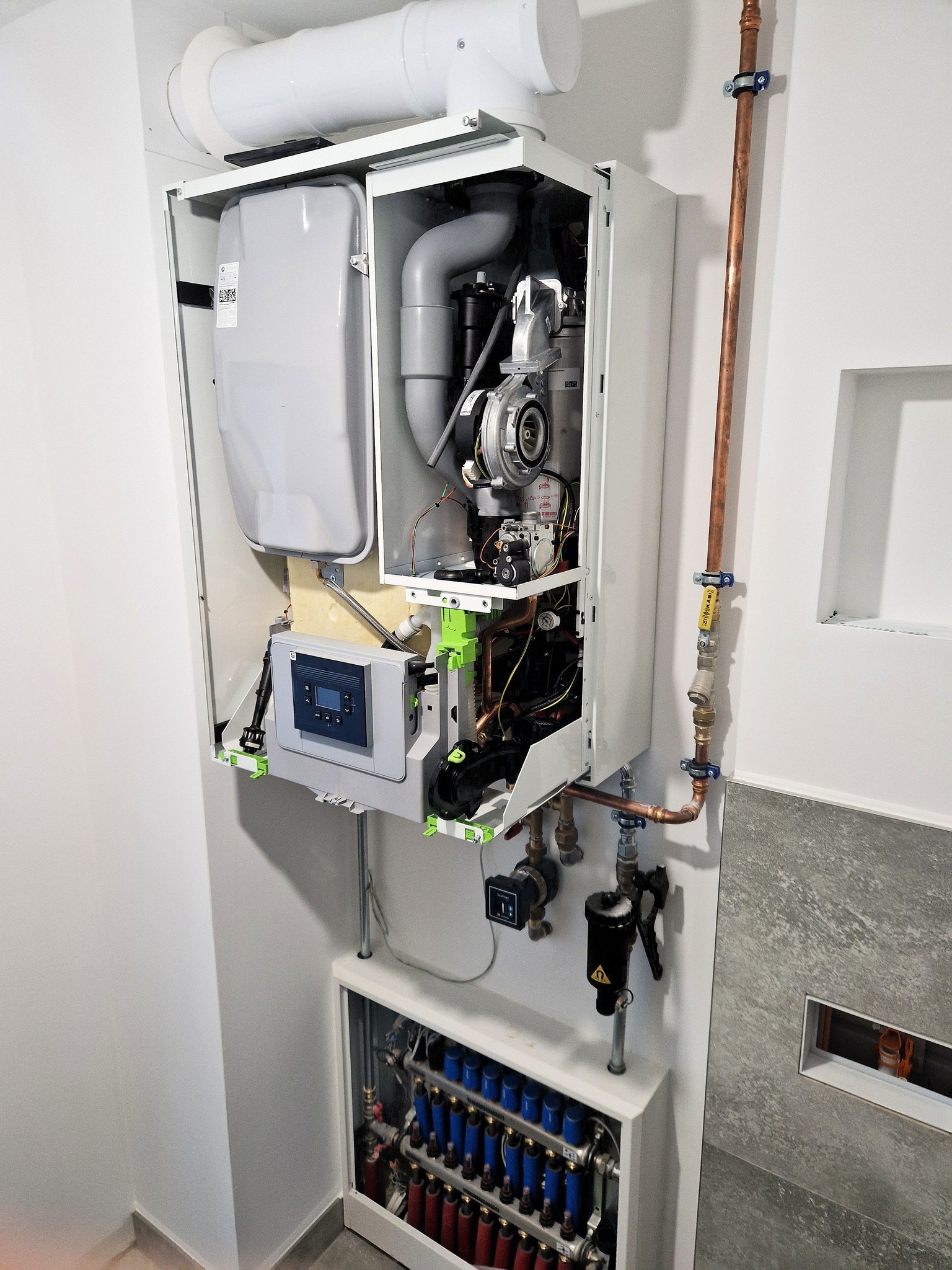 Nowoczesny piec gazowy z otwartą obudową, widoczne elementy instalacji CO i ogrzewania podłogowego, rury miedziane, białe ściany, szara płytka imitująca kamień.