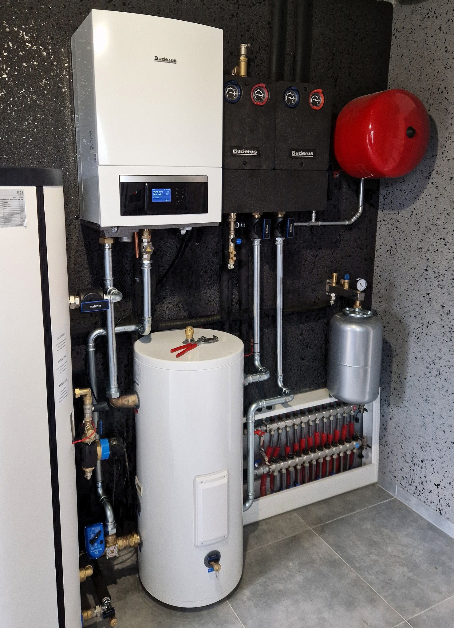 Nowoczesna kotłownia z piecem gazowym Buderus, zbiornikiem, naczyniem wzbiorczym i rozdzielaczem ogrzewania podłogowego na tle ściany o fakturze baranka.