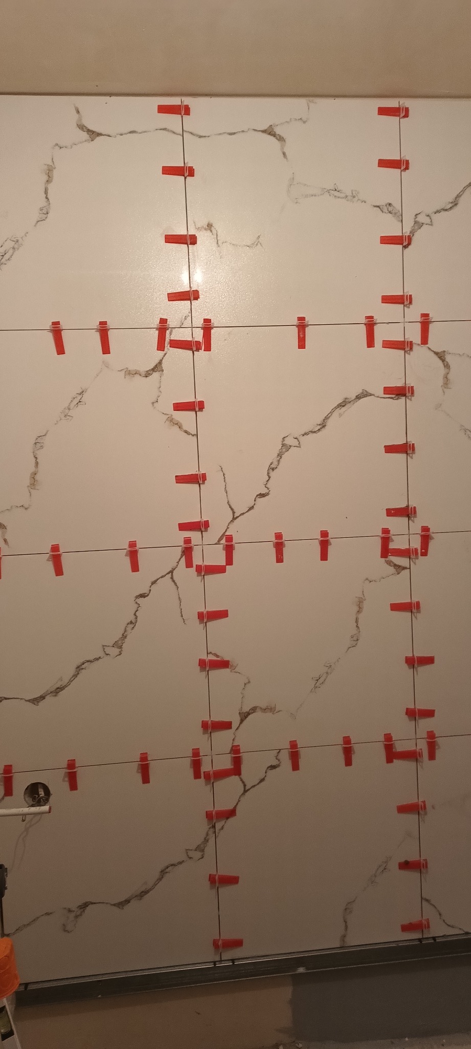 Ściana z białych płytek imitujących marmur, w trakcie montażu, z użyciem czerwonych klipsów poziomujących. Widoczne fugi i fragment podłogi.