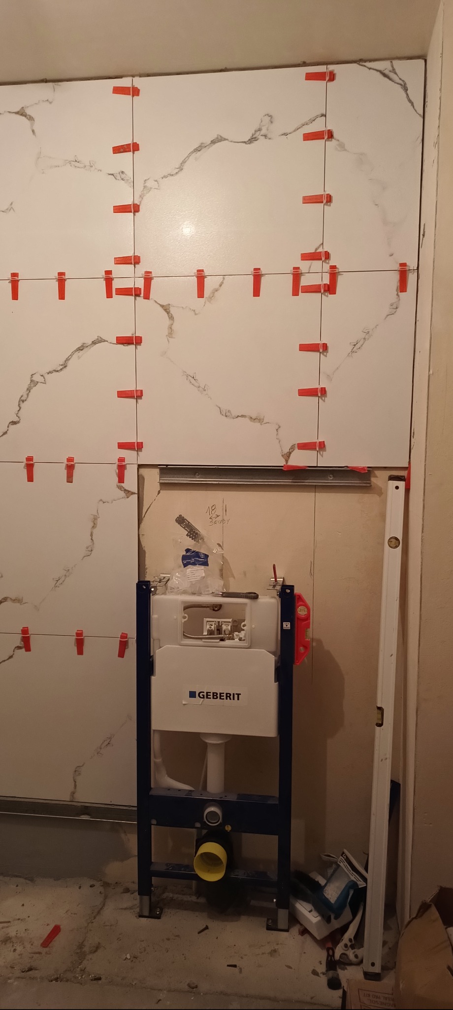Montaż płytek imitujących marmur w łazience, z użyciem czerwonych krzyżyków dystansowych. Widoczny stelaż podtynkowy WC Geberit i poziomica.