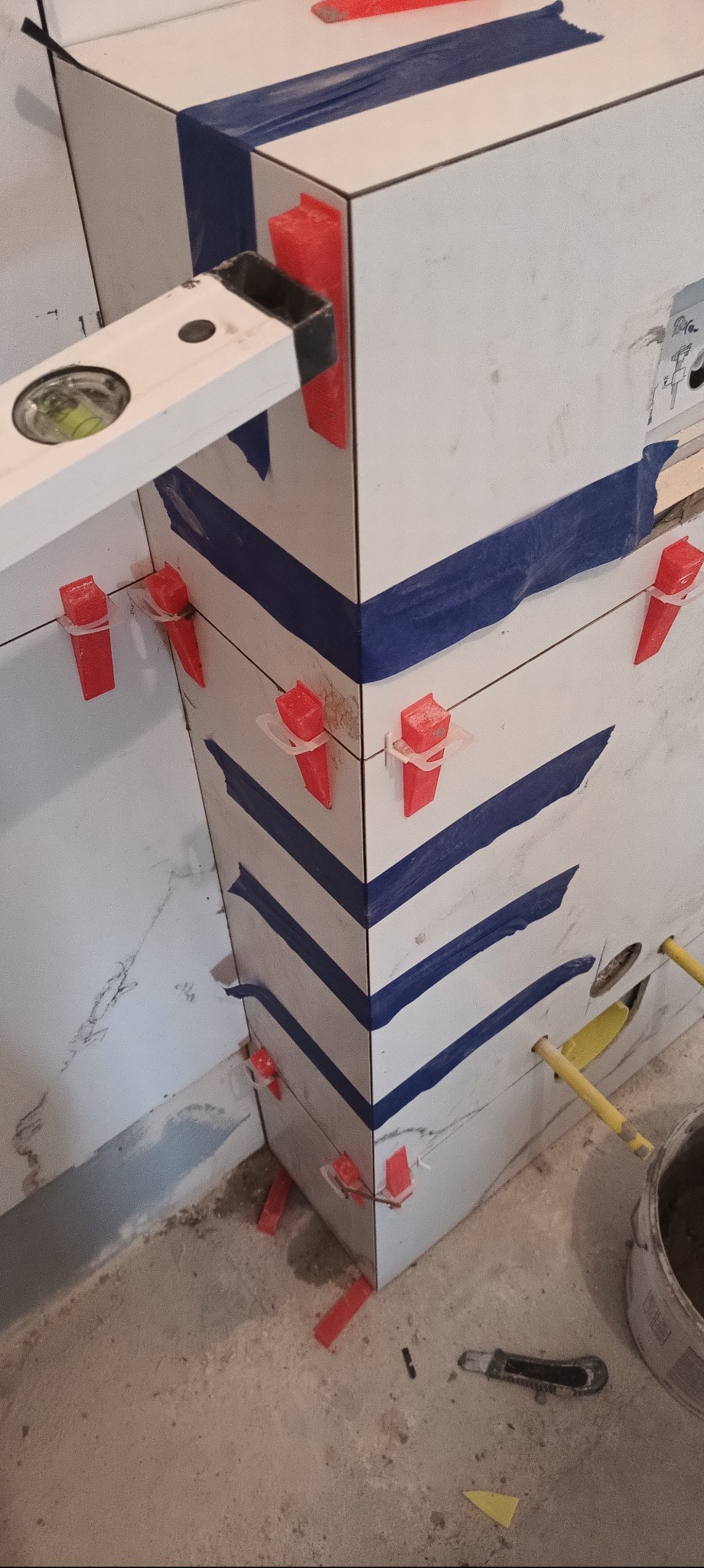 Układanie białych płytek z marmurowym wzorem, zabezpieczonych niebieską taśmą i czerwonymi klinami. Widoczna poziomica i wiadro z zaprawą.