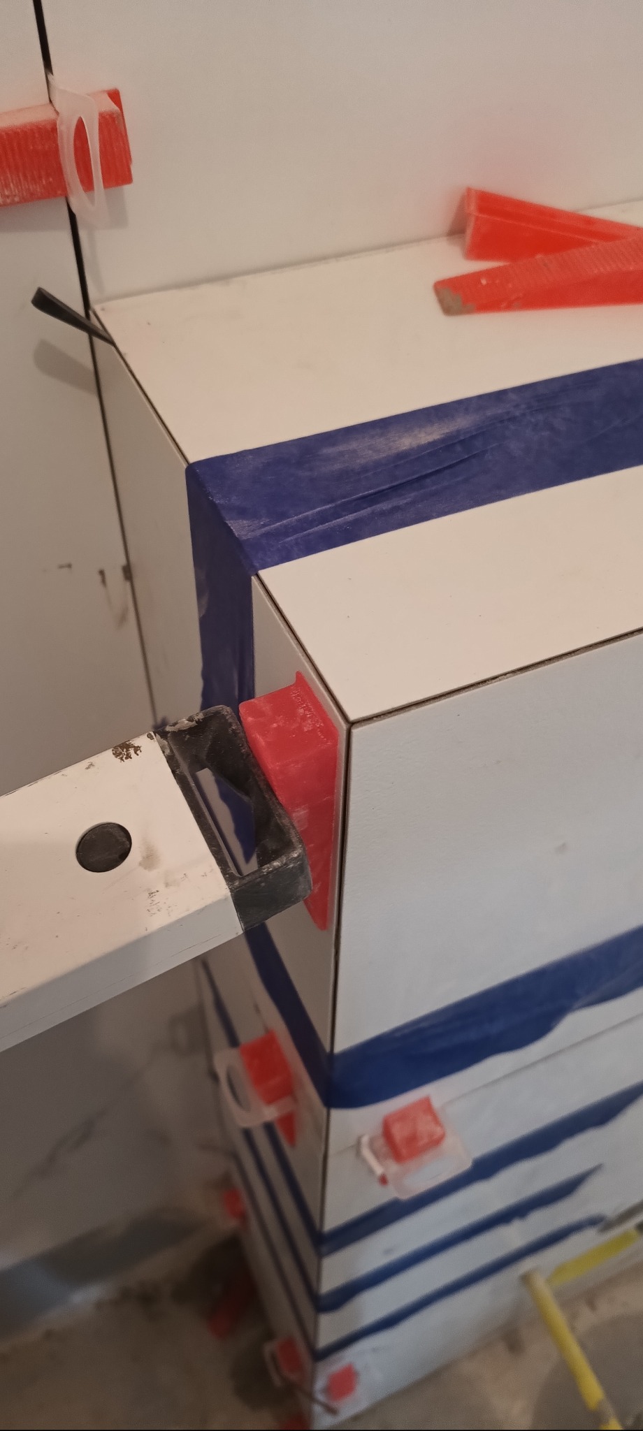 Układanie białych płytek z niebieskimi pasami, z użyciem poziomicy i czerwonych klinów dystansowych. Widoczne taśmy maskujące. Detal z bliska.