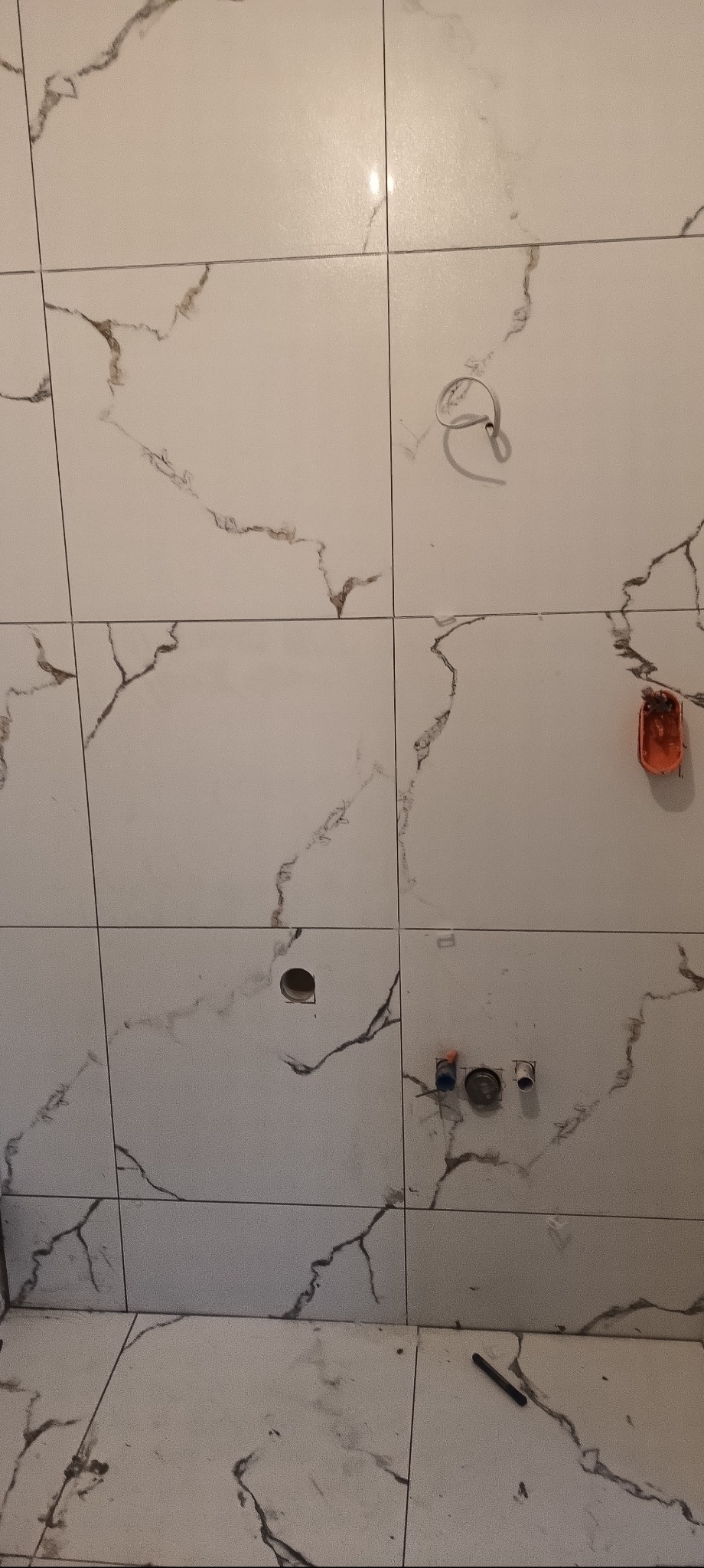 Ściana i podłoga wyłożone białymi płytkami imitującymi marmur, z widocznymi otworami na instalacje i wystającą puszką elektryczną. Na podłodze leży czarny pisak.