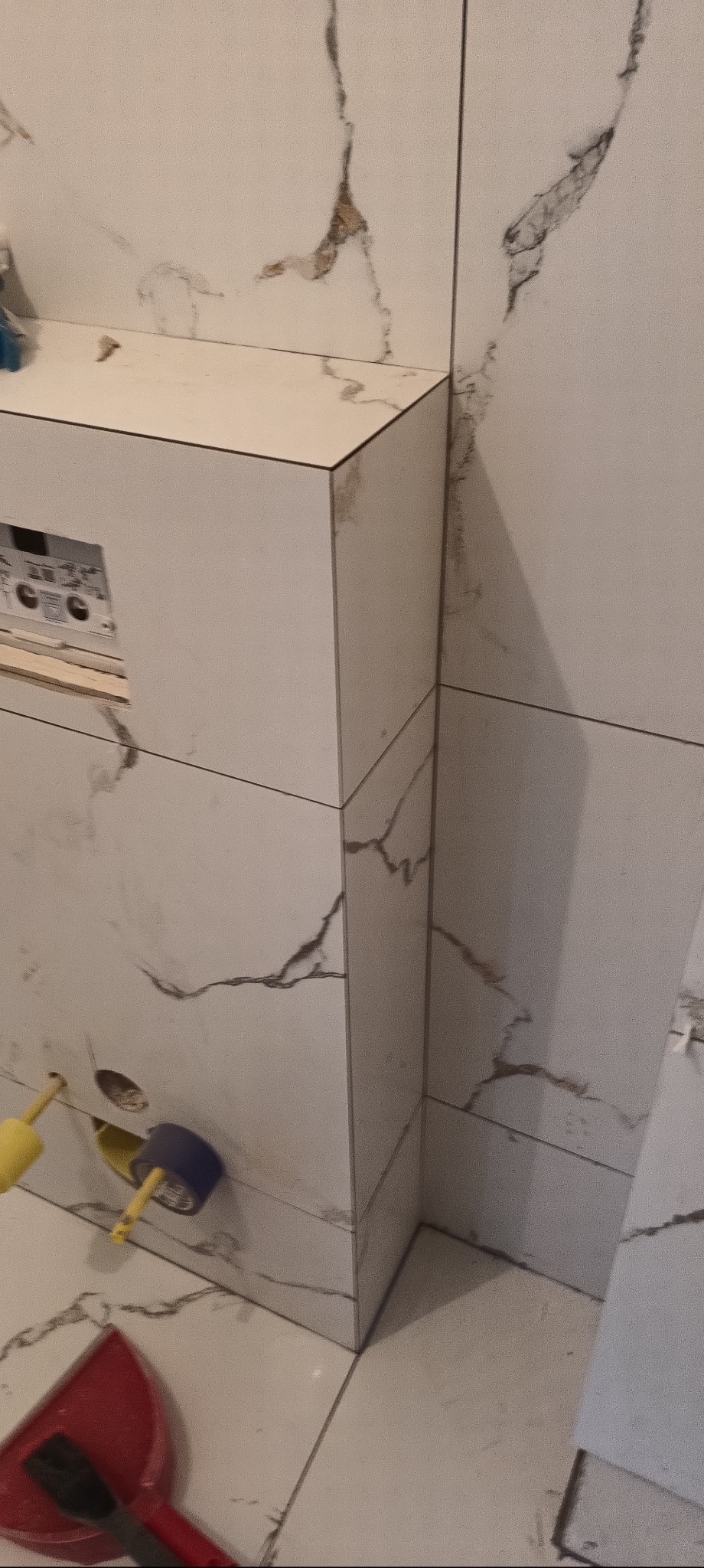 Wykończona obudowa WC z białych płytek imitujących marmur, widoczne narzędzia glazurnicze i otwór na przyciski spłukiwania. Minimalistyczny design łazienki.