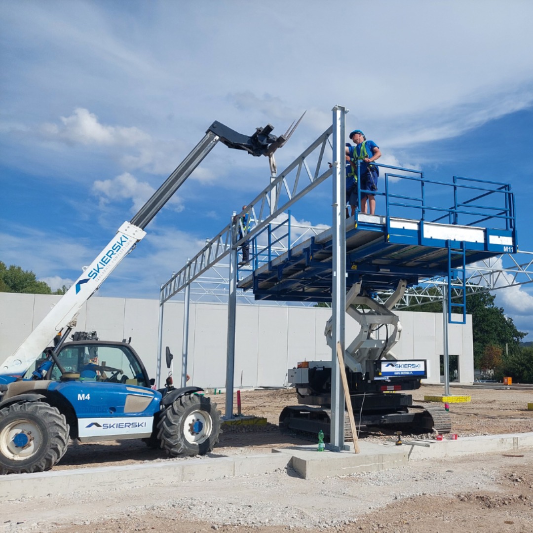 Montaż stalowej konstrukcji hali z użyciem podnośników teleskopowych i platform roboczych na tle błękitnego nieba z chmurami.