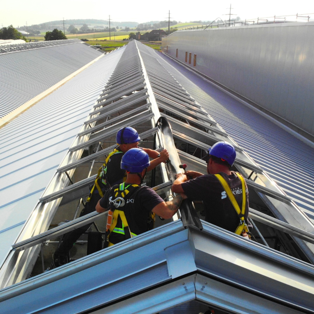 Trzech monterów w kaskach instaluje płytę na dachu hali z metalową konstrukcją. Widok z góry, w tle budynki i zielone pola.