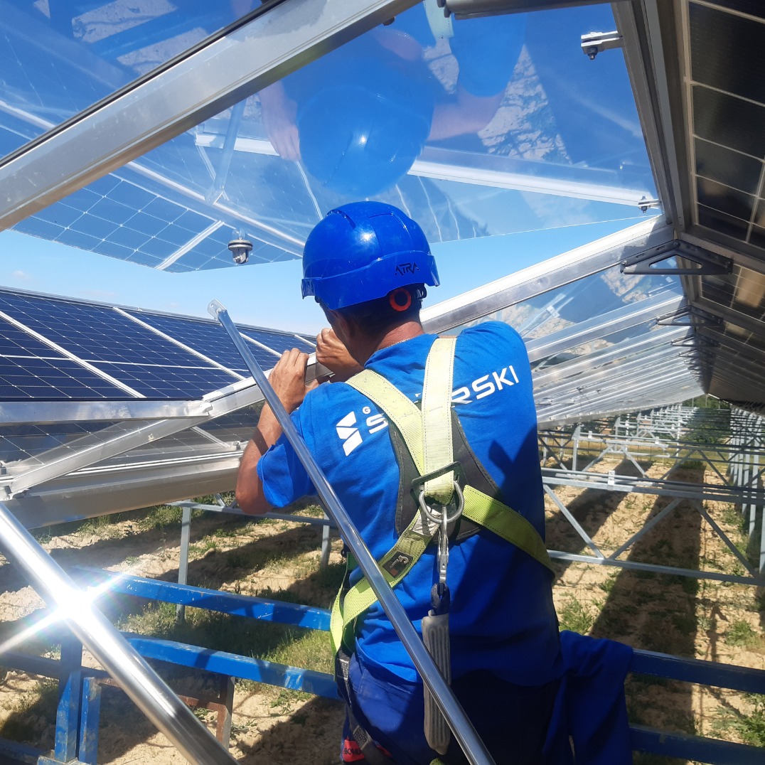 Pracownik w kasku i uprzęży montuje panele słoneczne na konstrukcji stalowej hali, widok z dołu, błękitne niebo w tle.