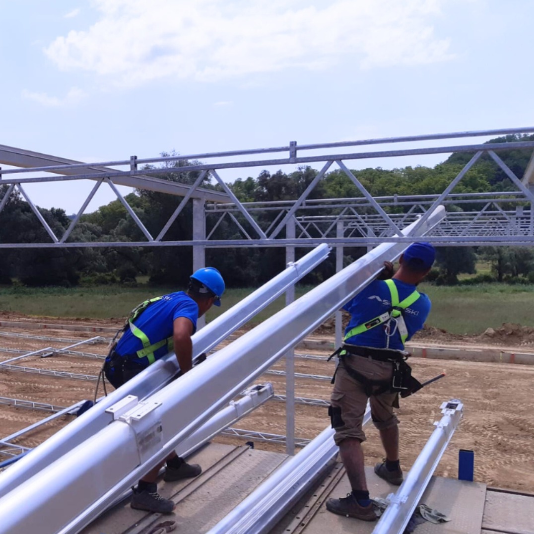 Dwóch monterów w niebieskich koszulkach i kaskach przenosi stalowe elementy konstrukcji hali na tle częściowo zmontowanego szkieletu i zieleni.