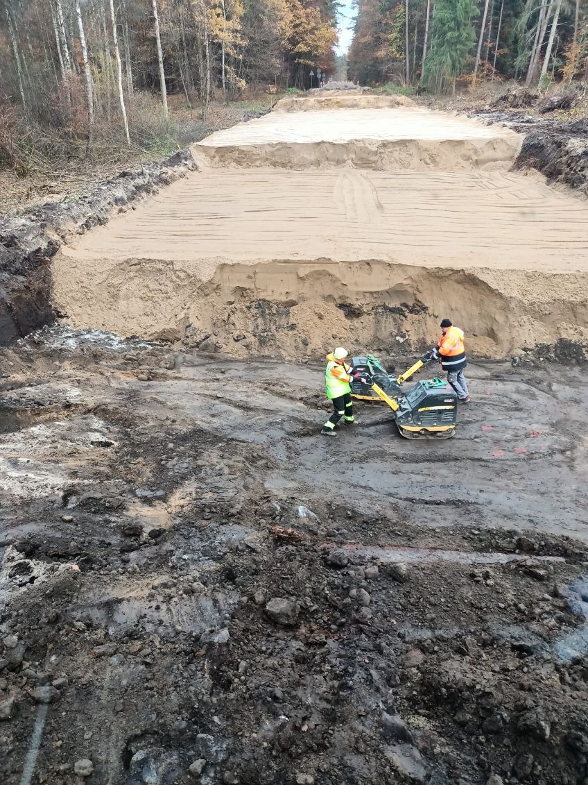 Dwaj pracownicy w odblaskowych kamizelkach obsługują zagęszczarki na błotnistym terenie, w tle wyrównany teren pod budowę drogi, otoczony lasem.
