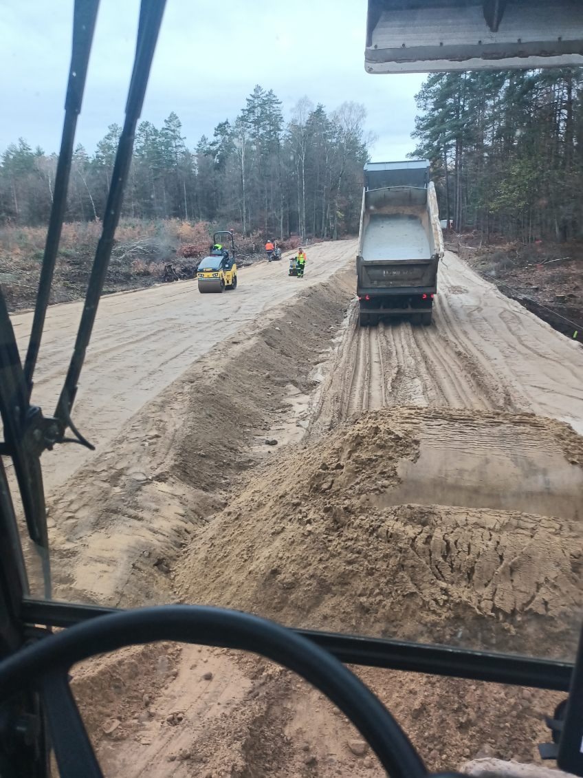 Widok z kabiny na utwardzanie drogi: wywrotka z piaskiem, walec i pracownicy w odblaskowych kamizelkach na tle lasu. Etap prac ziemnych.