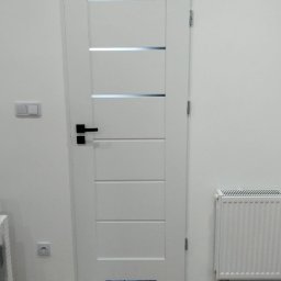USŁUGI REMONTOWO BUDOWLANE - Białe drzwi z czarną klamką i poziomymi przeszkleniami, zamontowane w jasnym wnętrzu z drewnianą podłogą i grzejnikiem. Minimalistyczny design.