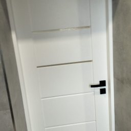 USŁUGI REMONTOWO BUDOWLANE - Białe, nowoczesne drzwi wewnętrzne z czarną klamką, zamontowane w łazience wykończonej szarą płytką. Minimalistyczny design.