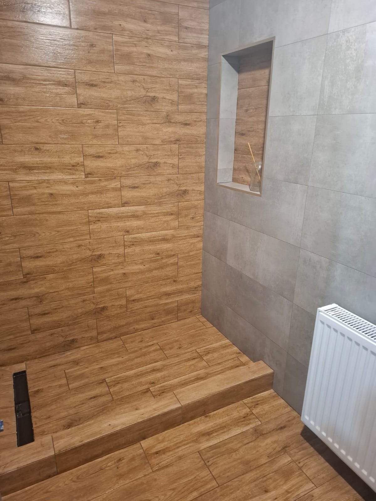 Wykończona łazienka z drewnopodobnymi i szarymi płytkami, widoczny grzejnik i wnęka w ścianie z dekoracją. Ujęcie z niskiego kąta podkreśla fakturę płytek.