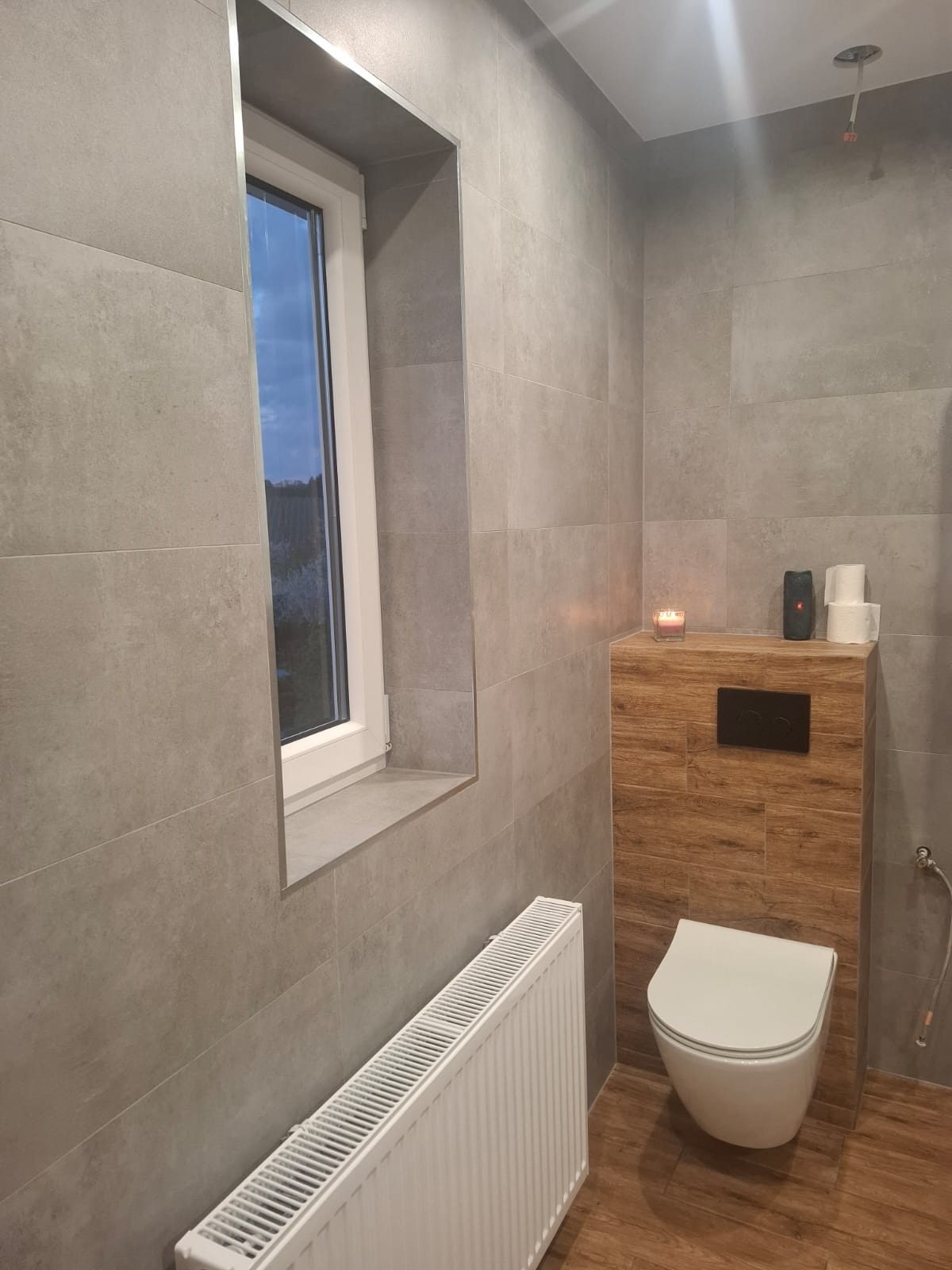 Nowoczesna łazienka z szarymi płytkami, podwieszaną toaletą i drewnianą zabudową. Okno z głębokim parapetem, biały grzejnik. Minimalistyczny design.