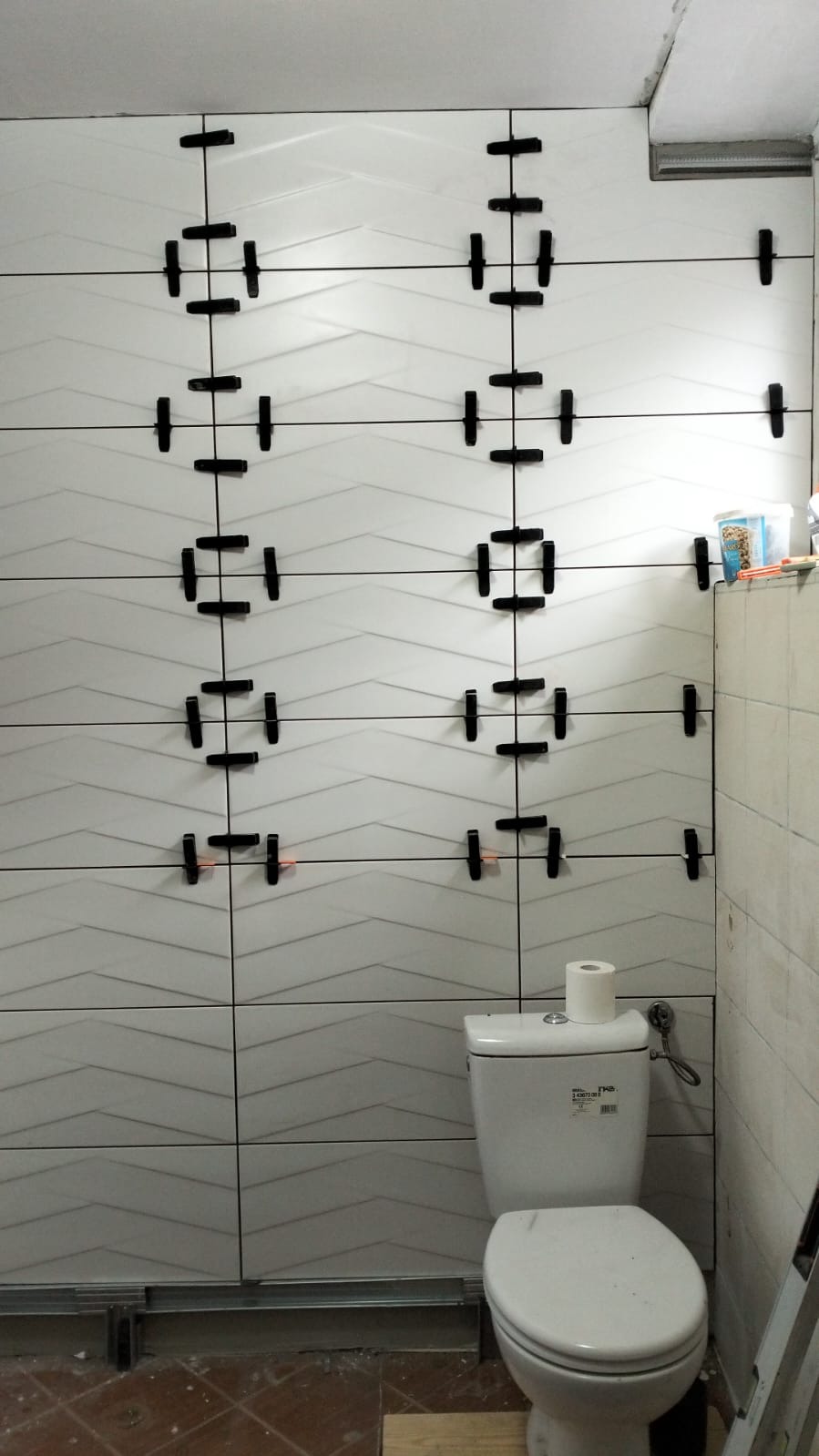 Białe, faliste płytki ceramiczne na ścianie łazienki, w trakcie montażu, z użyciem czarnych krzyżyków dystansowych. Widoczna toaleta i fragment podłogi.