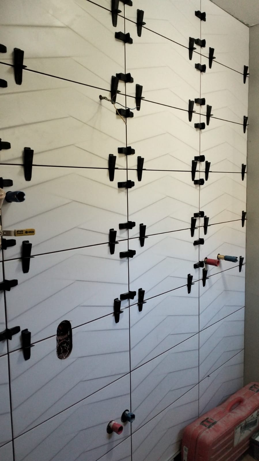 Ściana z białych, wzorzystych płytek w trakcie układania, z użyciem czarnych krzyżyków dystansowych i systemu poziomowania. Widoczne otwory na instalacje.