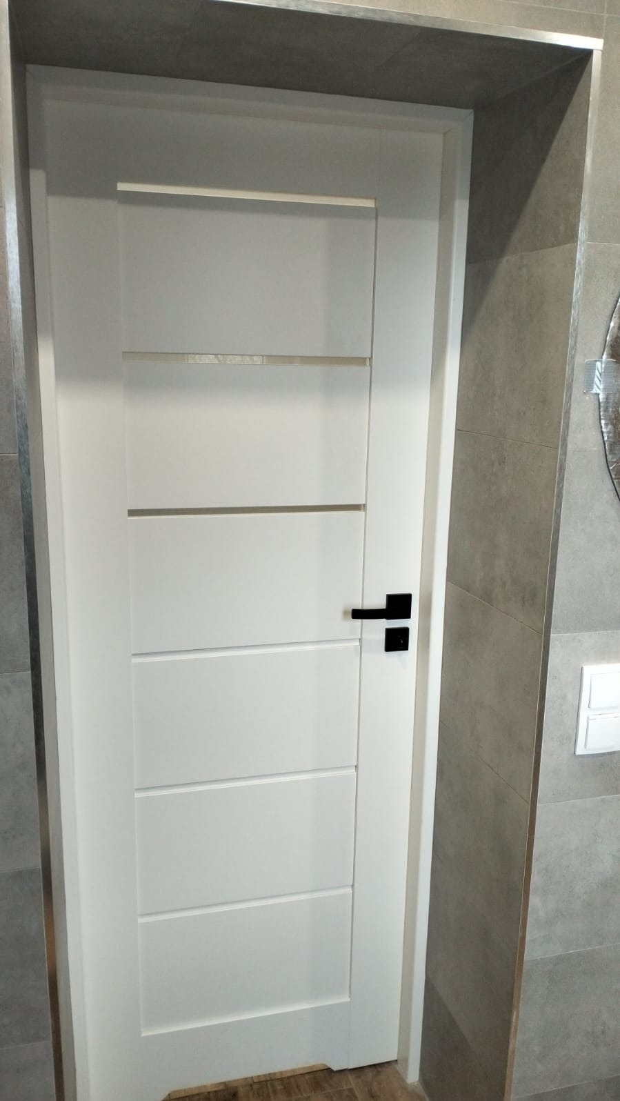 Białe, nowoczesne drzwi wewnętrzne z czarną klamką, zamontowane w łazience wykończonej szarą płytką. Widoczny fragment lustra i włącznik światła.