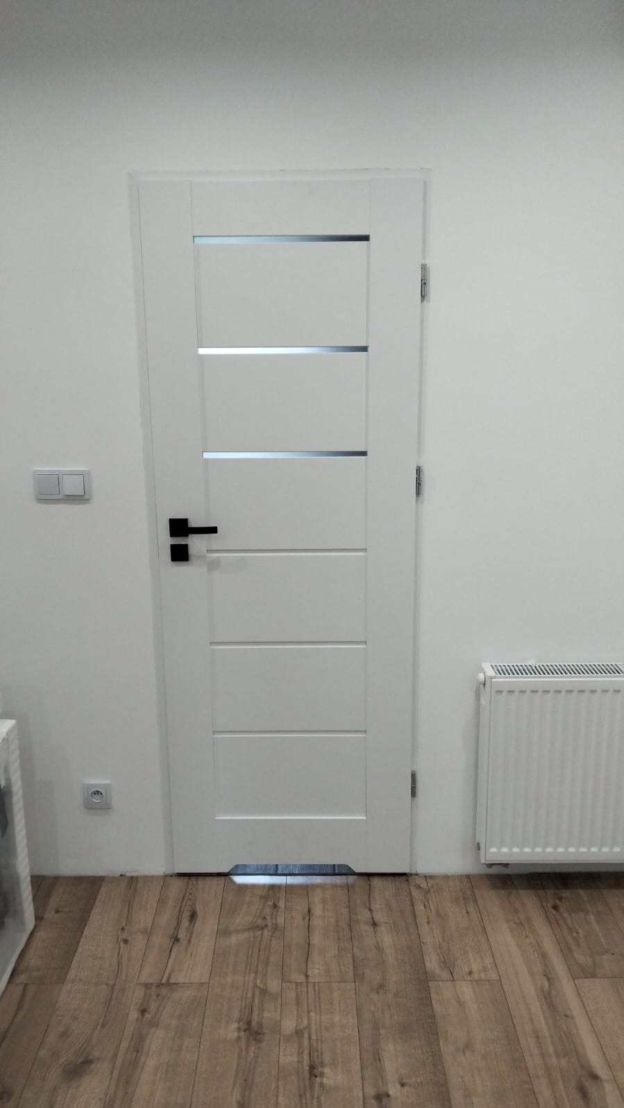 Białe drzwi z czarną klamką i poziomymi przeszkleniami, zamontowane w jasnym wnętrzu z drewnianą podłogą i grzejnikiem. Minimalistyczny design.