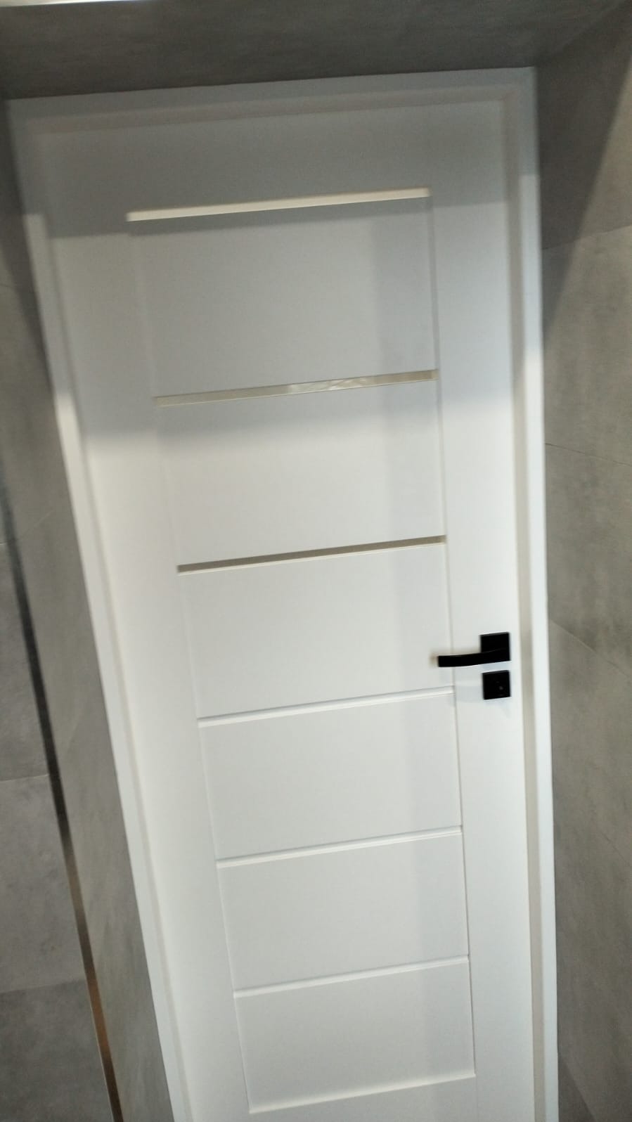 Białe, nowoczesne drzwi wewnętrzne z czarną klamką, zamontowane w łazience wykończonej szarą płytką. Minimalistyczny design.