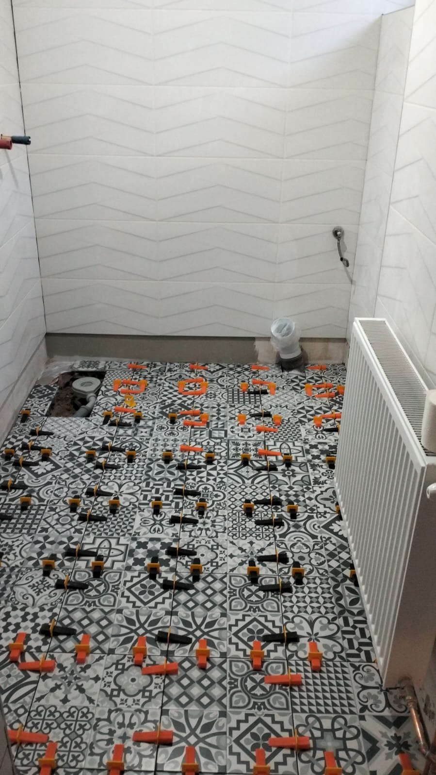 Układanie wzorzystych płytek podłogowych w łazience z użyciem klipsów poziomujących. Widoczne białe płytki ścienne z delikatnym wzorem i grzejnik.