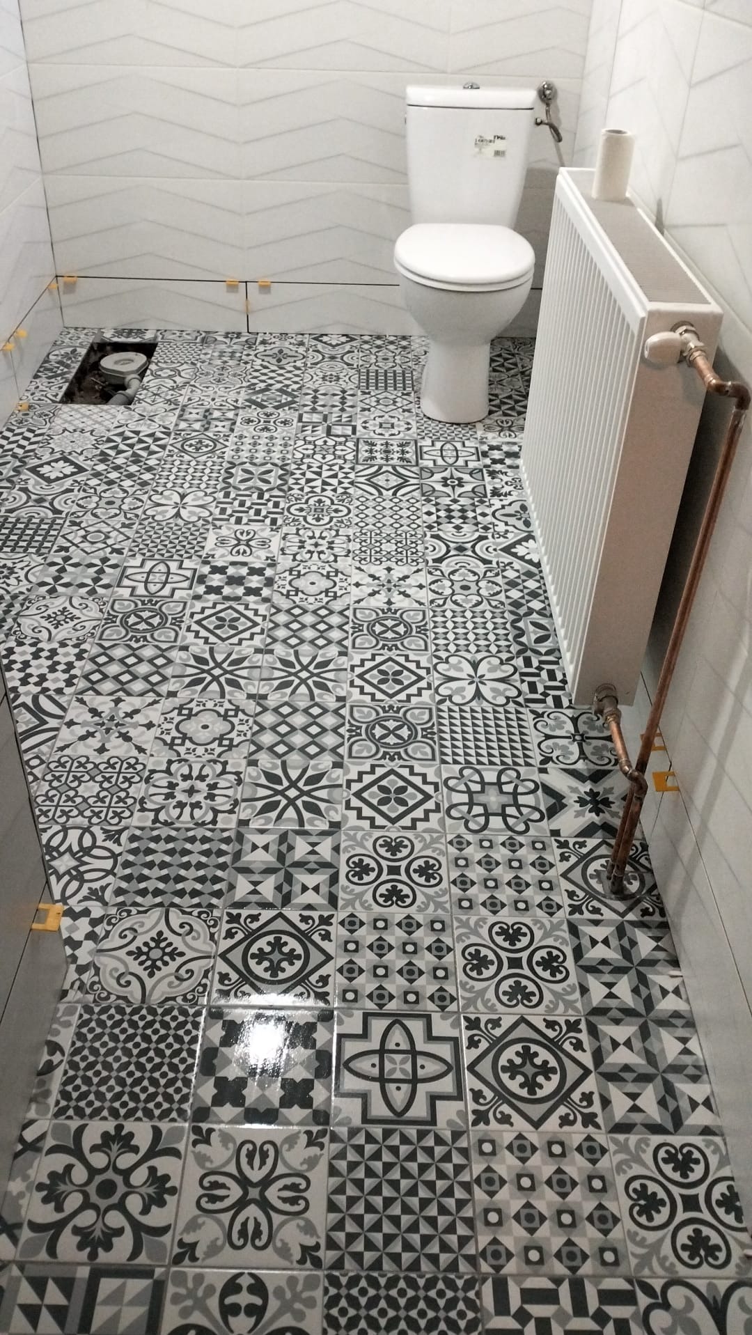 Ułożona podłoga z wzorzystych, czarno-białych płytek w łazience. Widoczny fragment toalety, grzejnik i otwór w podłodze. Żółte krzyżyki dystansowe między płytkami.