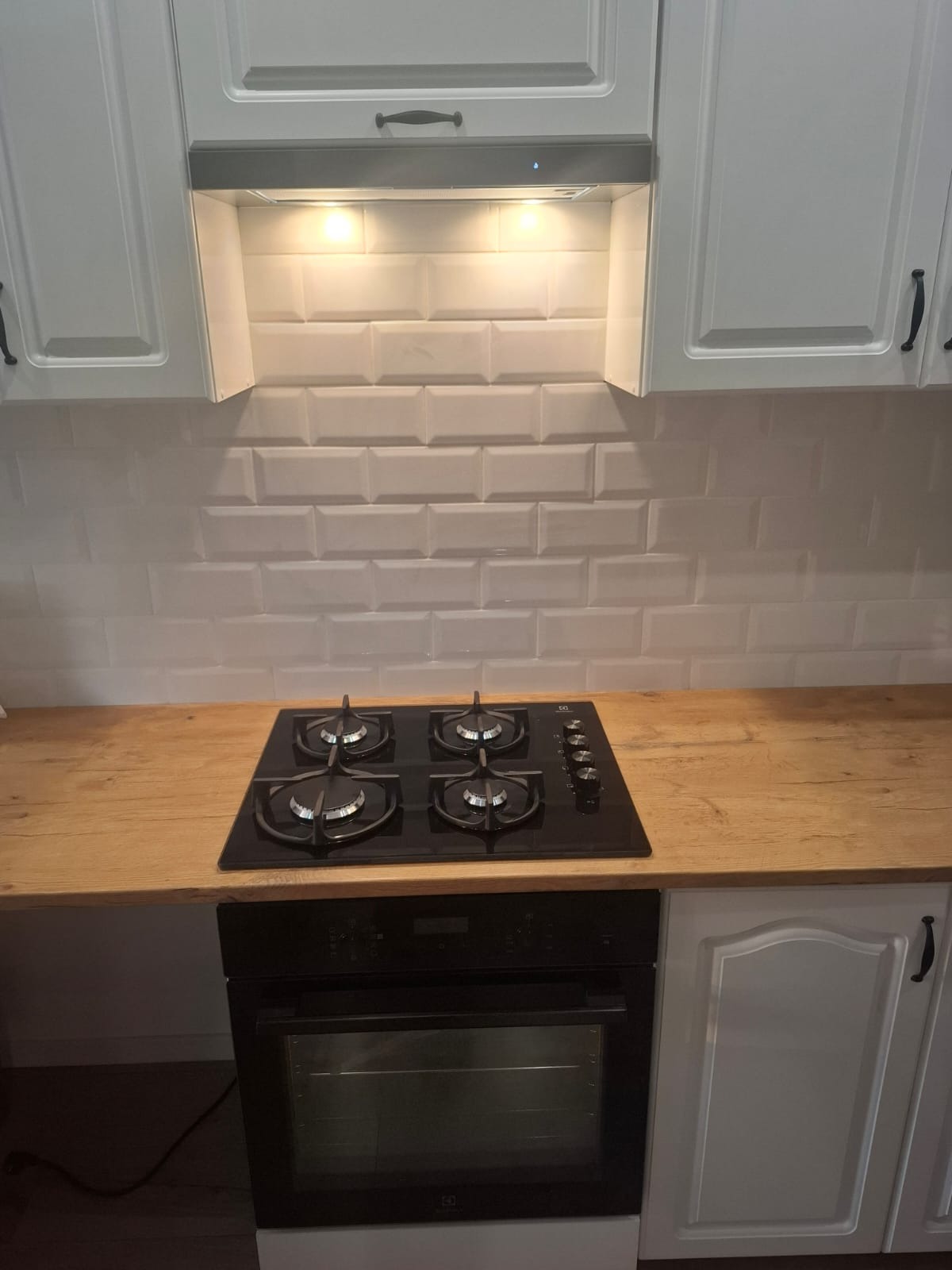 Biała kuchnia z drewnianym blatem i płytkami typu metro. Czarna płyta gazowa i piekarnik. Ujęcie na wykończoną zabudowę kuchenną z oświetleniem podszafkowym.