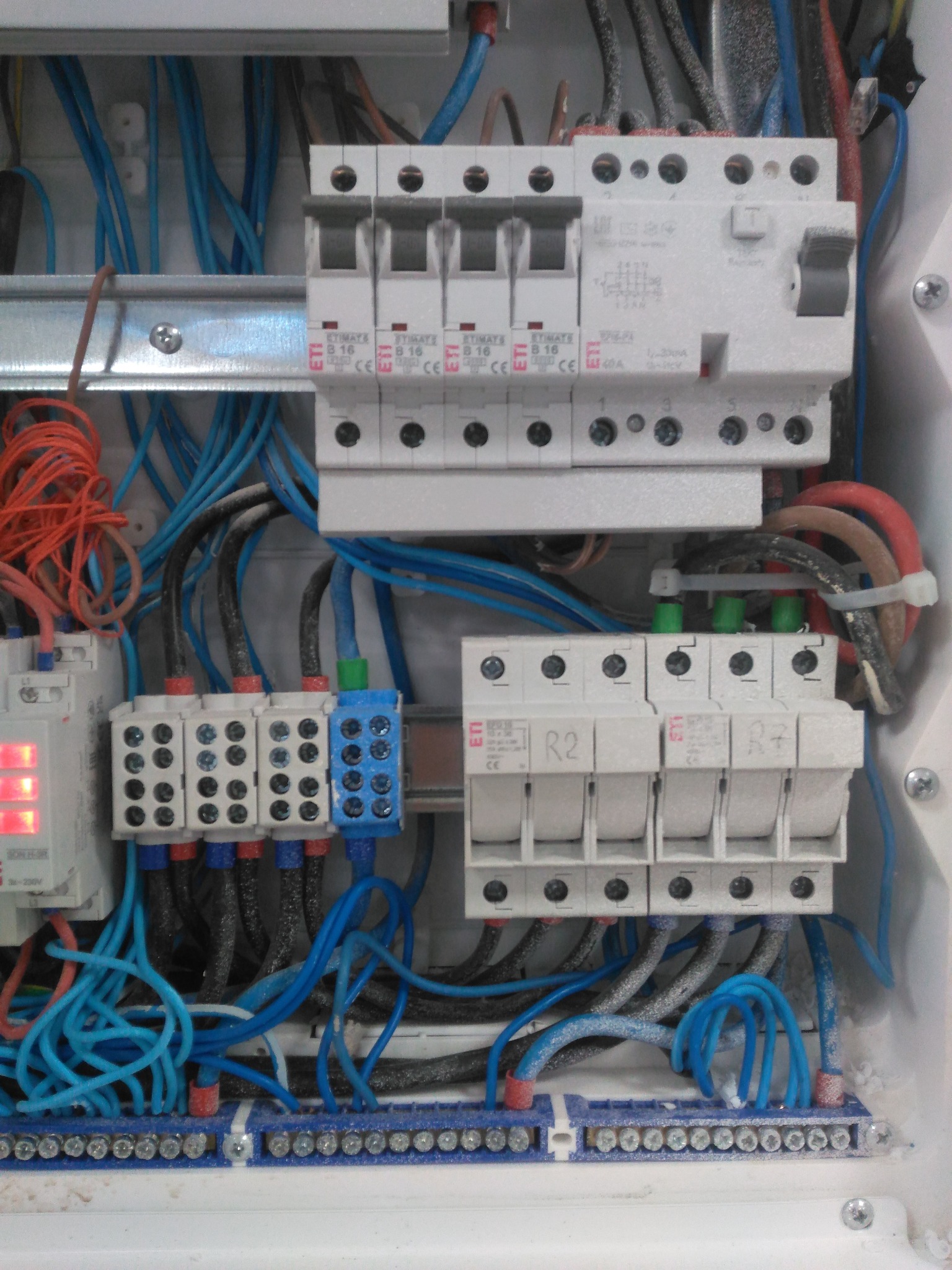 Rozdzielnia elektryczna z bezpiecznikami, przewodami i listwami zaciskowymi wewnątrz białej obudowy. Widoczne oznaczenia na elementach i różnokolorowe przewody.