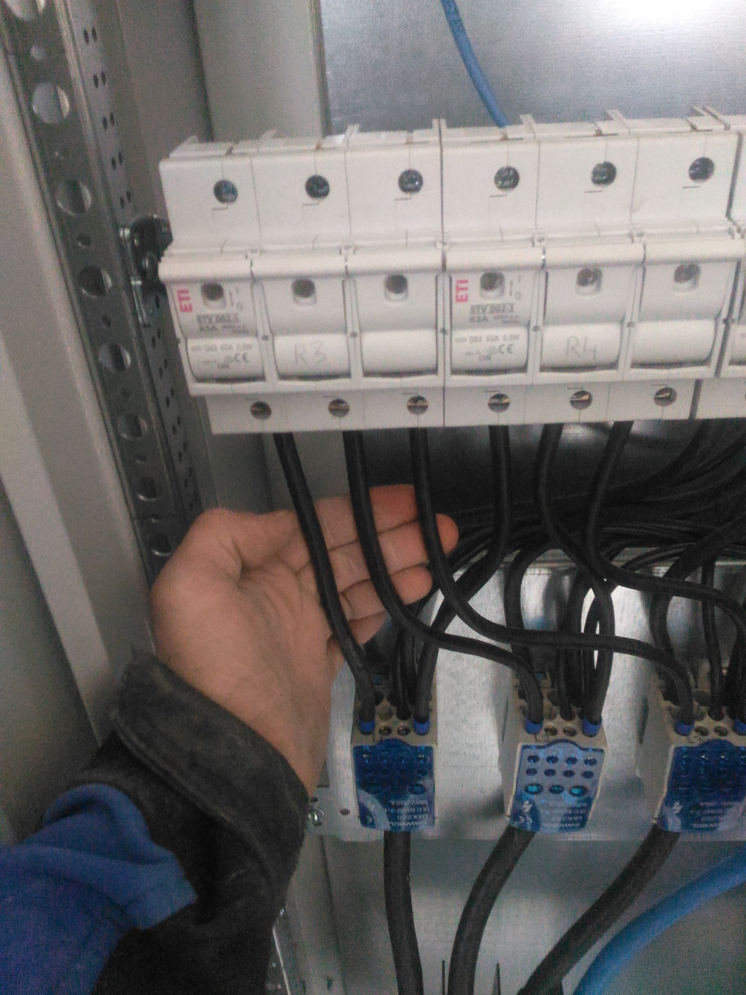 Ręka elektryka przy bezpiecznikach i przewodach w metalowej szafie rozdzielczej. Widoczne oznaczenia R3 i R4 na bezpiecznikach. Przewody czarne i niebieskie.