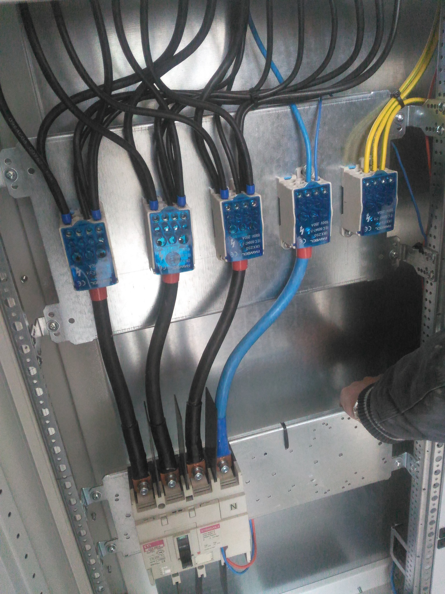 Rozdzielnica elektryczna z podłączonymi przewodami o różnych kolorach (czarny, niebieski, żółty) i zabezpieczeniami. Widoczne elementy montażowe i fragment dłoni.