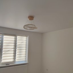 VOLODYMYR LUTSENKO - Jasne wnętrze po remoncie: świeżo pomalowane ściany w odcieniach bieli, okno z żaluzjami i ozdobny żyrandol. Podłoga przygotowana do dalszych prac wykończeniowych.