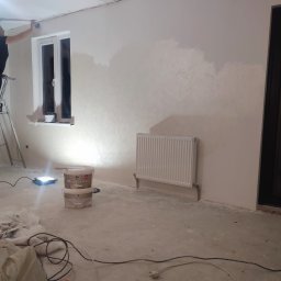 VOLODYMYR LUTSENKO - Wnętrze w trakcie malowania: ściana z grzejnikiem, okno, drabina i wiaderka z farbą Mega Latex. Widoczne niedomalowane fragmenty ściany i narzędzia malarskie.