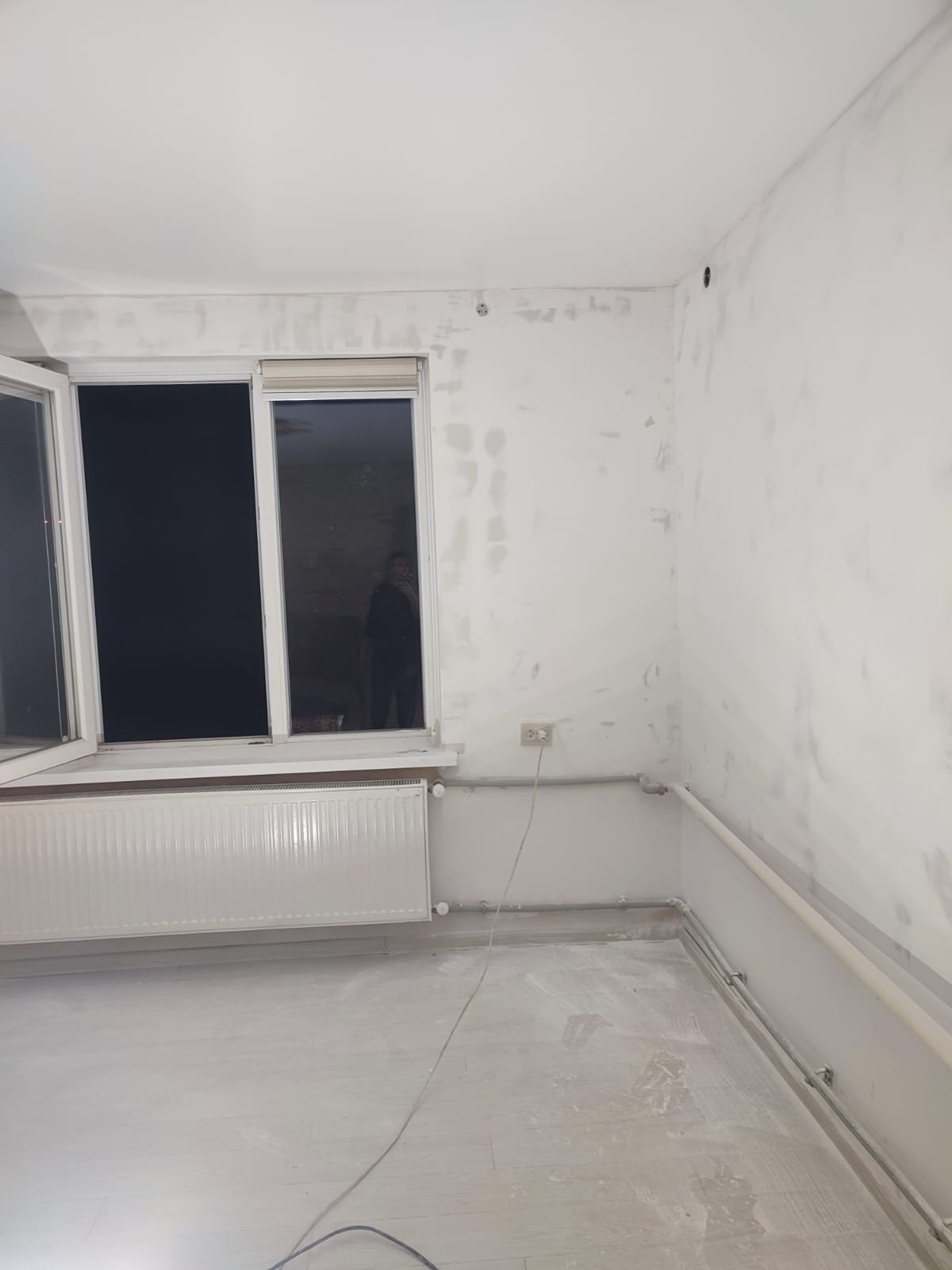 Pokój w trakcie przygotowania do malowania: ściany ze śladami szpachlowania, otwarte okno, grzejnik, podłoga zabezpieczona folią. Widoczne gniazdko elektryczne.