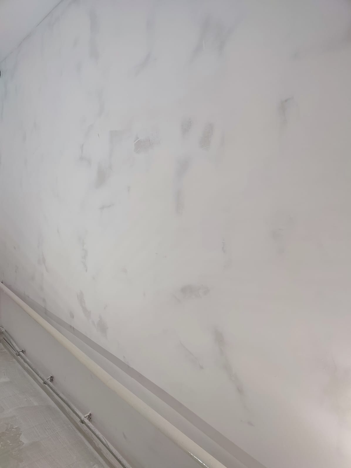 Ściana po szpachlowaniu z widocznymi smugami i rurami przy podłodze. Delikatne, szare odcienie na białym tle. Wykończenie gotowe do dalszych prac.